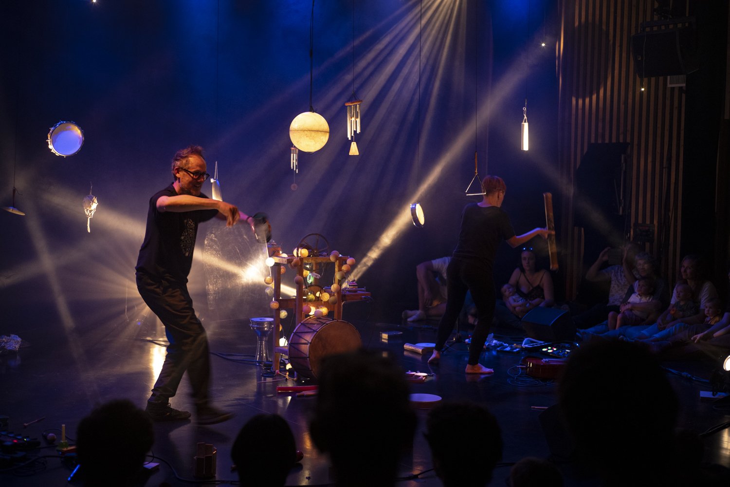 Spectacle de musique et d'animation avec deux artistes sur scène entourés de lumières et d'objets suspendus, avec un public assis sur le sol.