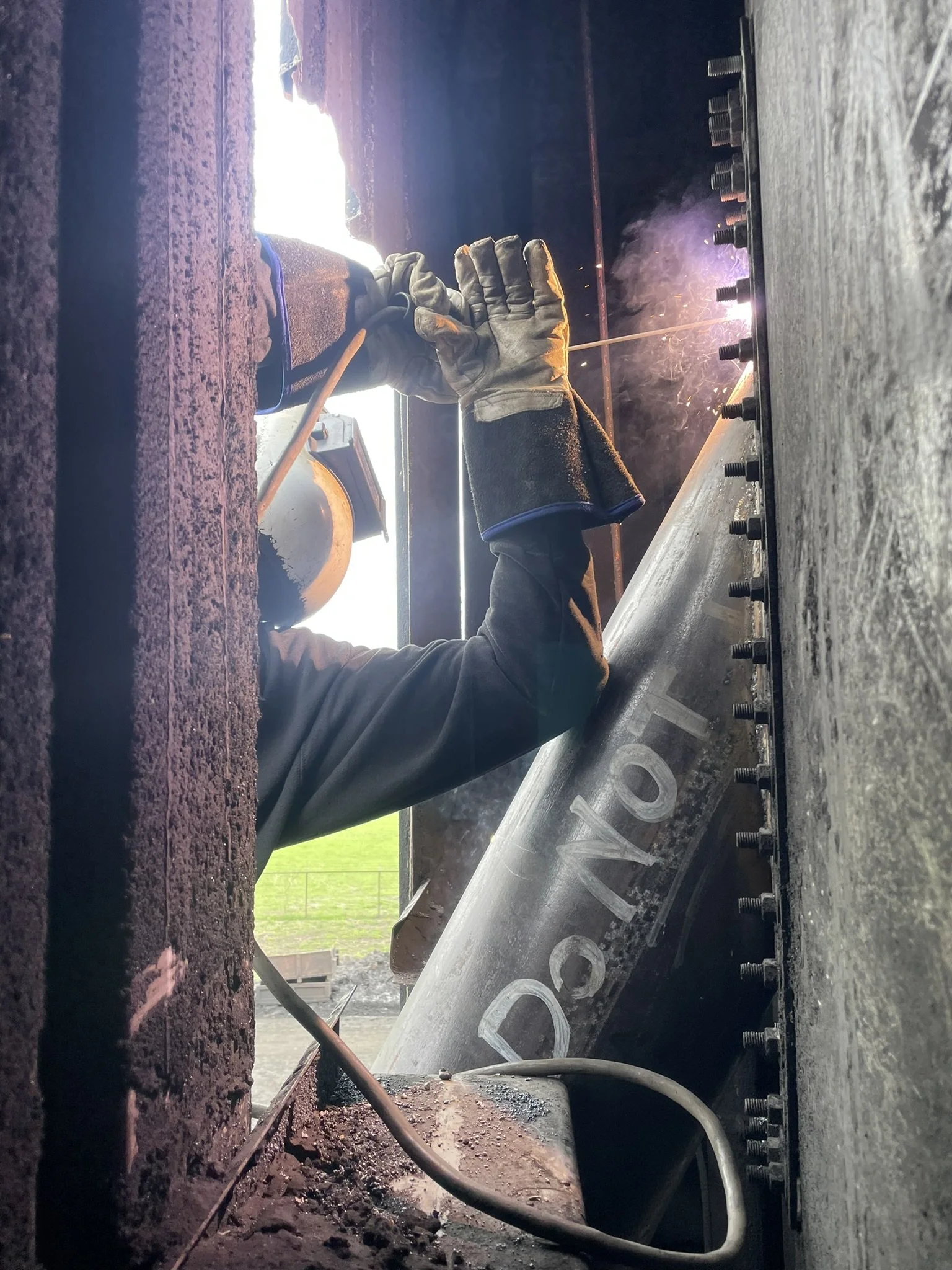 Welding process on a metal surface with sparks and smoke, closer view of a welder's gloved hand, welding tools, and metal parts in an industrial setting.