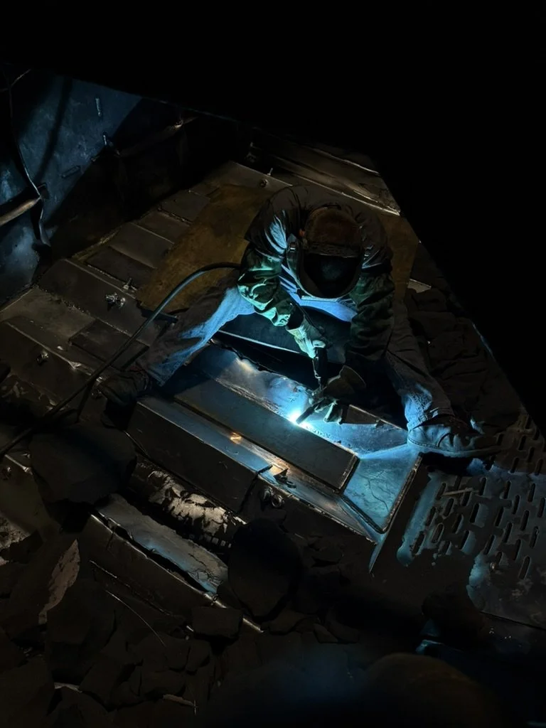 A person welding metal inside a dark industrial or workshop setting, with protective gloves and a hood, producing a bright blue welding arc.