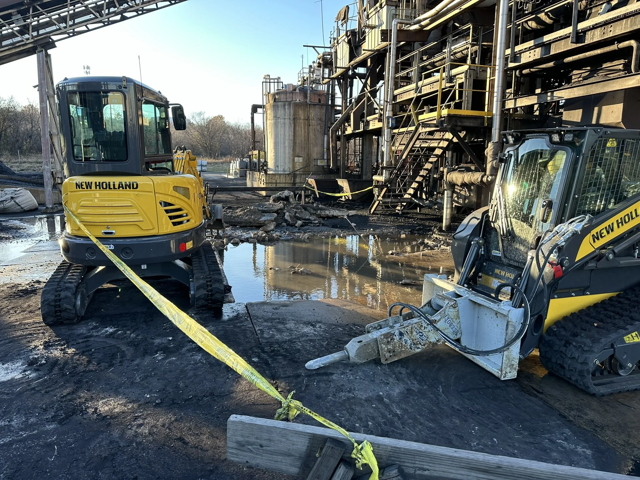 Construction site with yellow New Holland mini excavators, muddy ground, water puddles, industrial pipes, safety barriers, and a large rusted tank, under a clear sky.