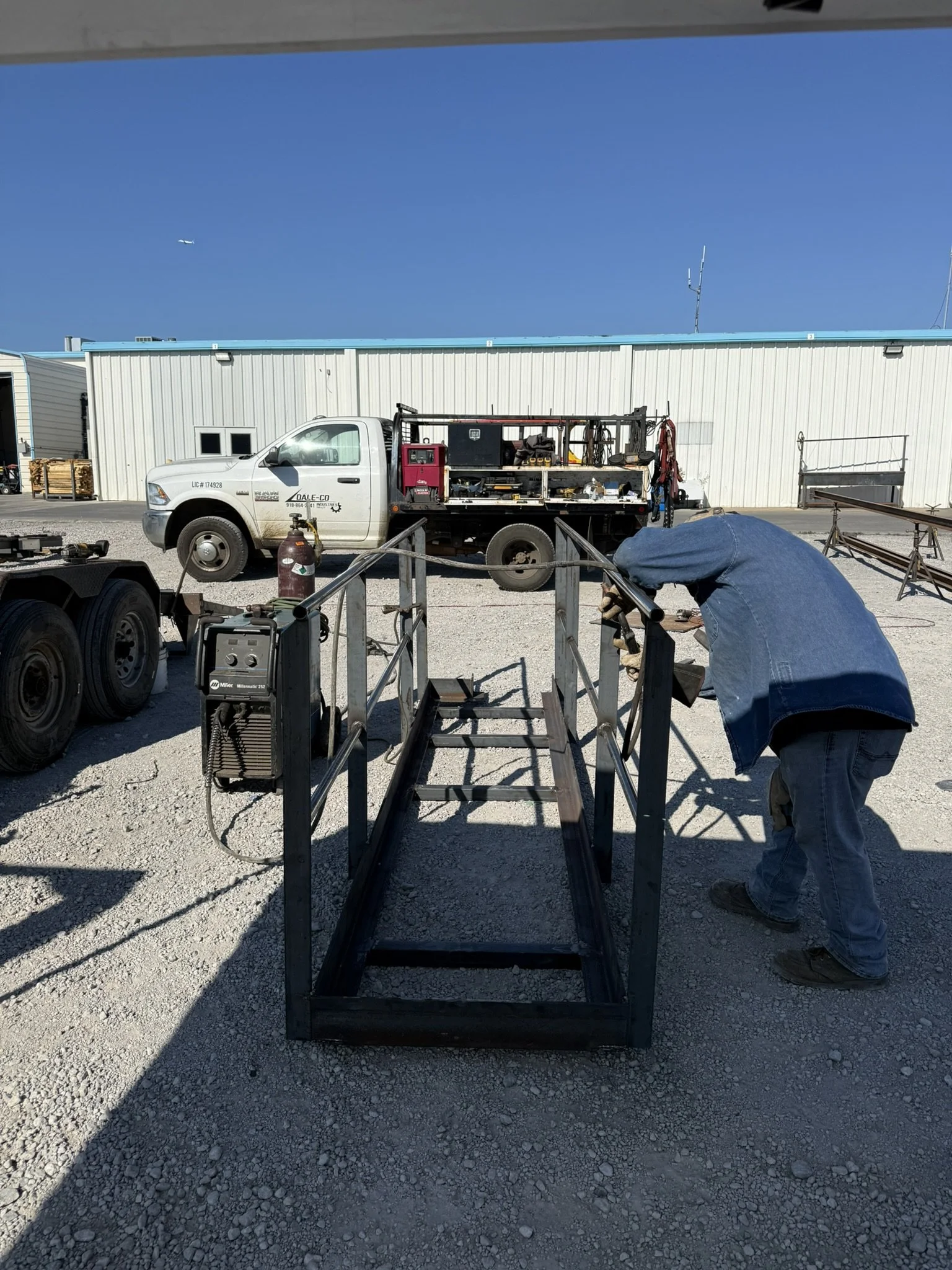 A worker in a blue jacket is welding metal in an outdoor industrial workspace. There is a construction truck with tools and equipment parked behind, and a white building with a blue roof in the background. The ground is gravel.