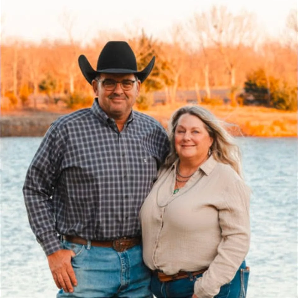 A man and woman standing close together outdoors by a body of water, with trees and a fence in the background during sunset. The man is wearing a cowboy hat, glasses, a plaid shirt, and jeans. The woman is wearing a light-colored shirt and jeans.