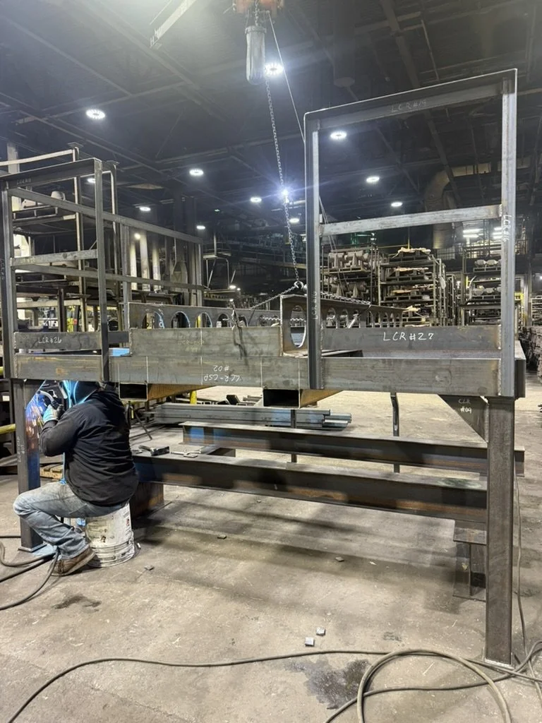 A worker welding a metal structure in an industrial factory setting, with various metal frameworks and equipment in the background.