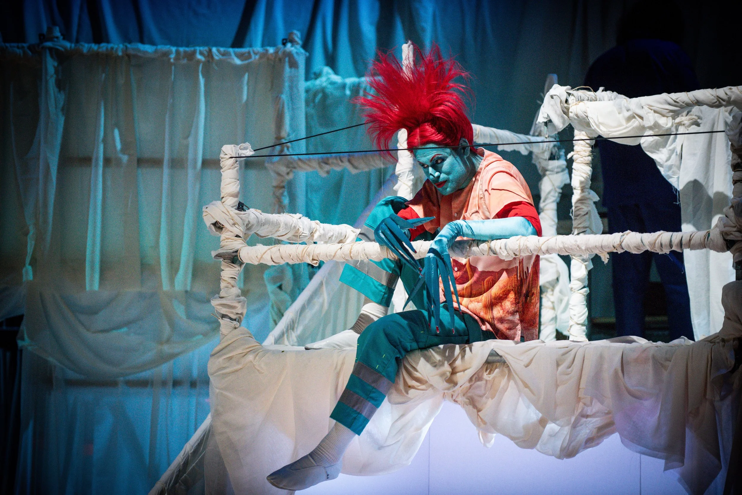 Clown met blauwe huid, fel rood haar en kleurrijk kostuum ziet op een telefoon terwijl ze op een decor met wit doek en touwen zit.