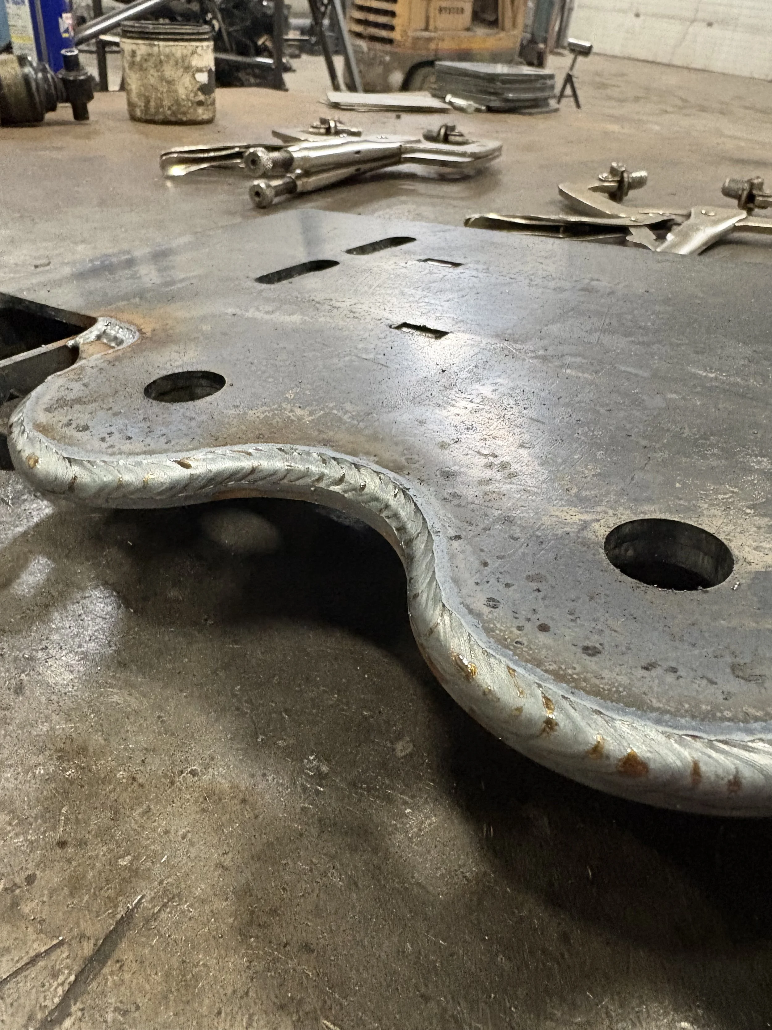 Close-up view of a metal car part on a workshop floor, with various other metallic tools and materials in the background.