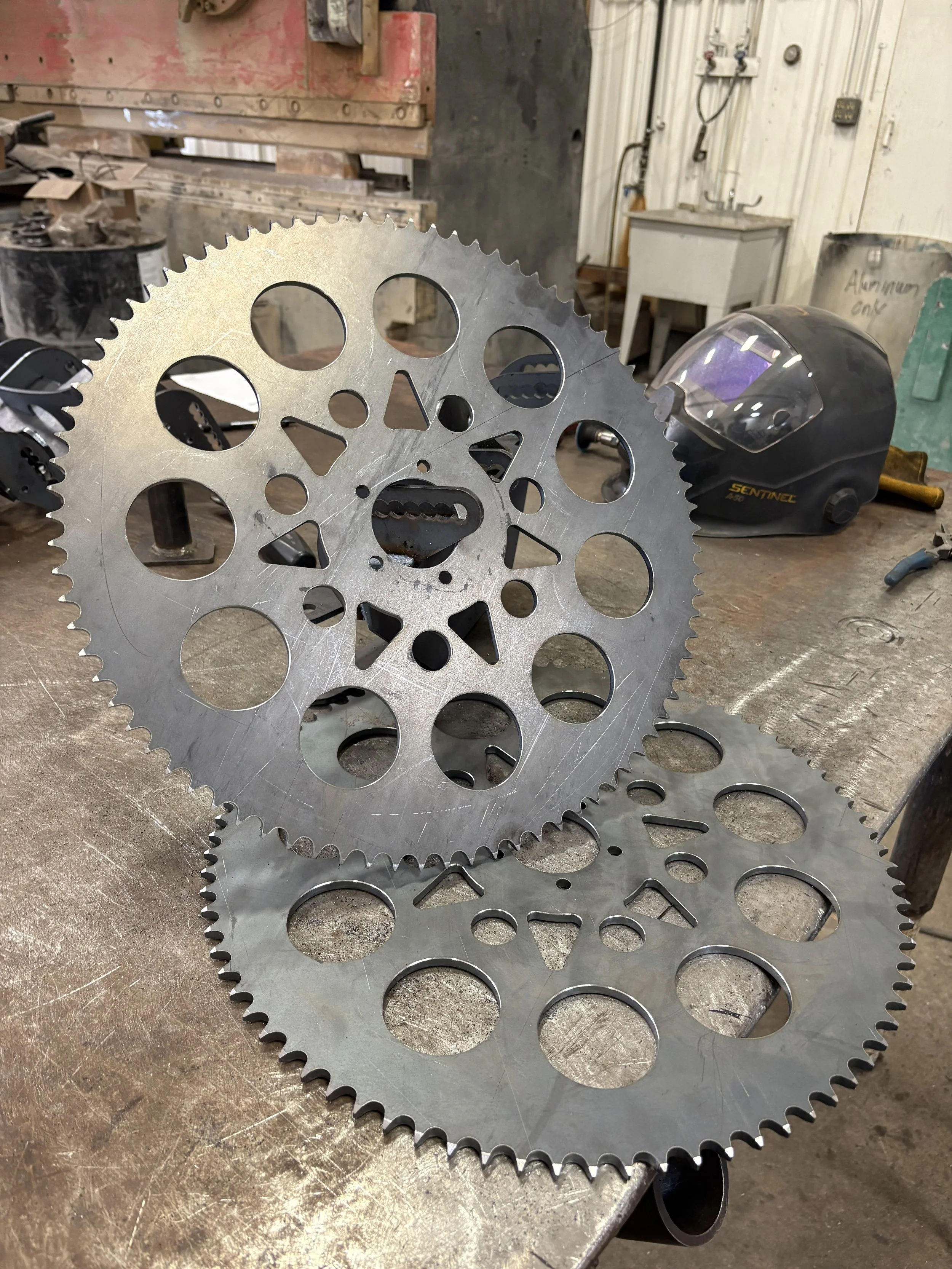 Two large metallic sprockets with circular and triangular cutouts on a workbench in an industrial or workshop setting, with welding helmet and tools nearby.