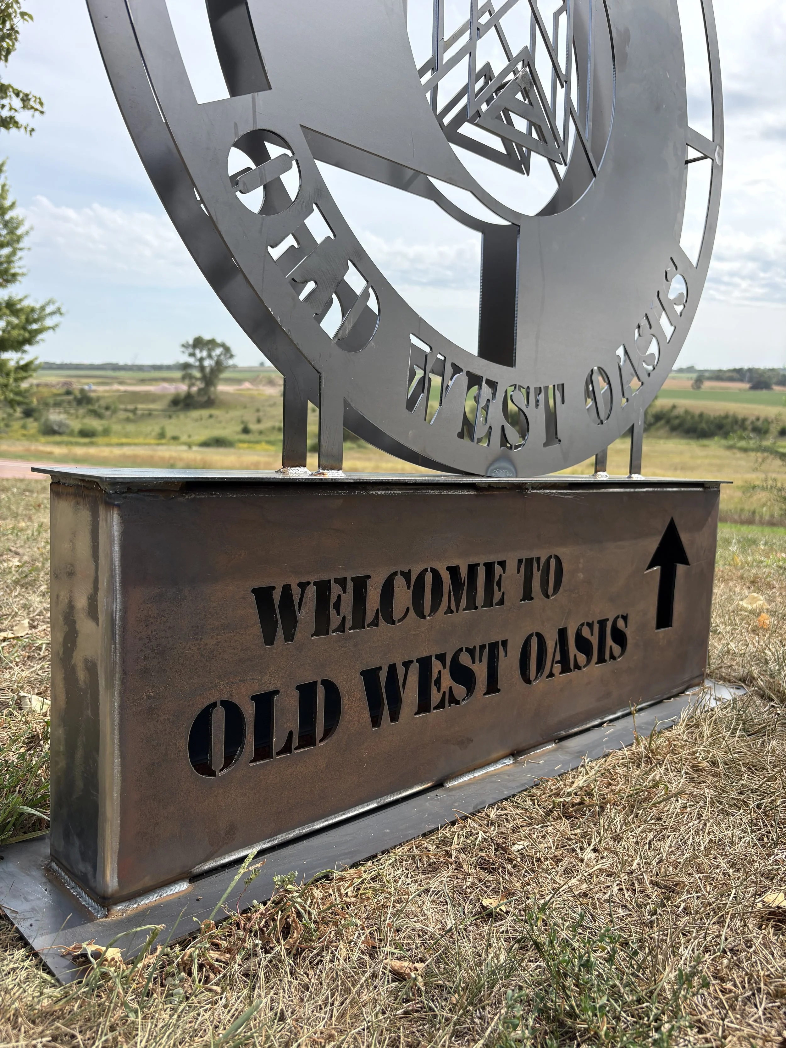 Metal sign with cut-out text reading 'Welcome to Old West Oasis' in a rural landscape with open fields, trees, and a partly cloudy sky.