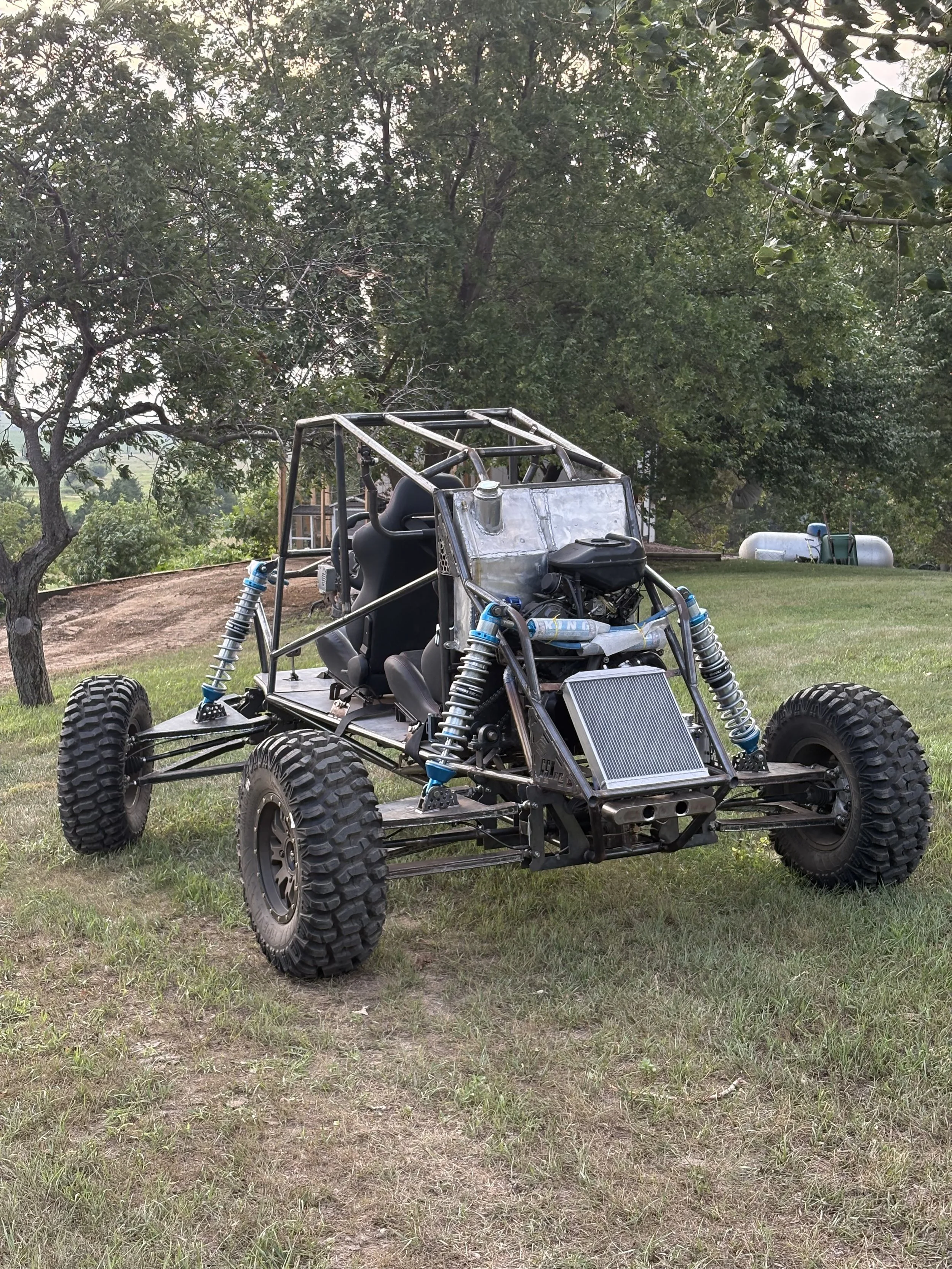 An assembled off-road vehicle with large tires, a roll cage, and exposed metal framework parked on grass with trees and a propane tank in the background.