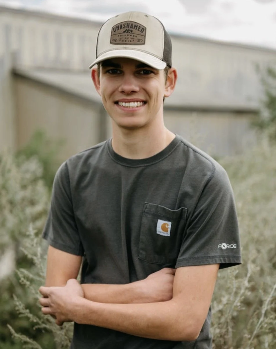 Cade Foster, the business owner, smiling outdoors wearing a beige and gray trucker hat with the word 'Unashamed' on it, and a dark gray Carhartt t-shirt with a chest pocket.