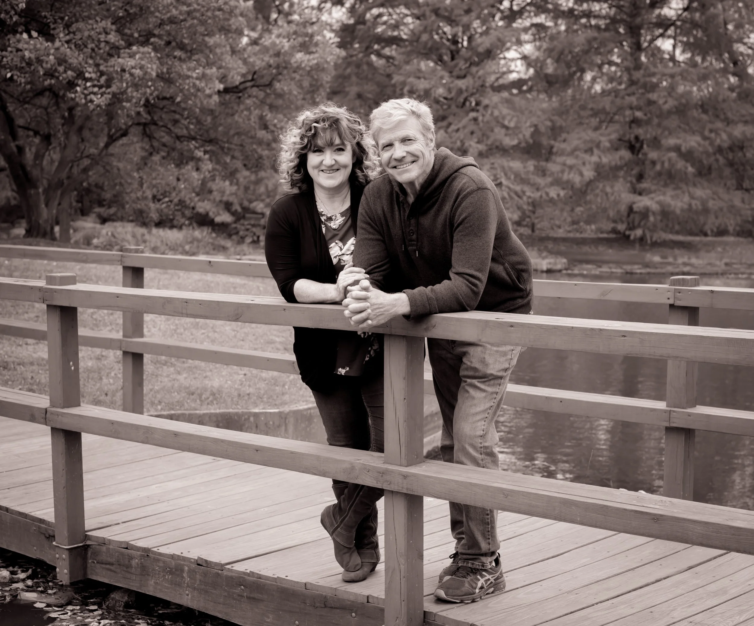 A smiling couple stands leaning on a wooden railing of a bridge or boardwalk outdoors in a park or nature setting, with trees in the background.