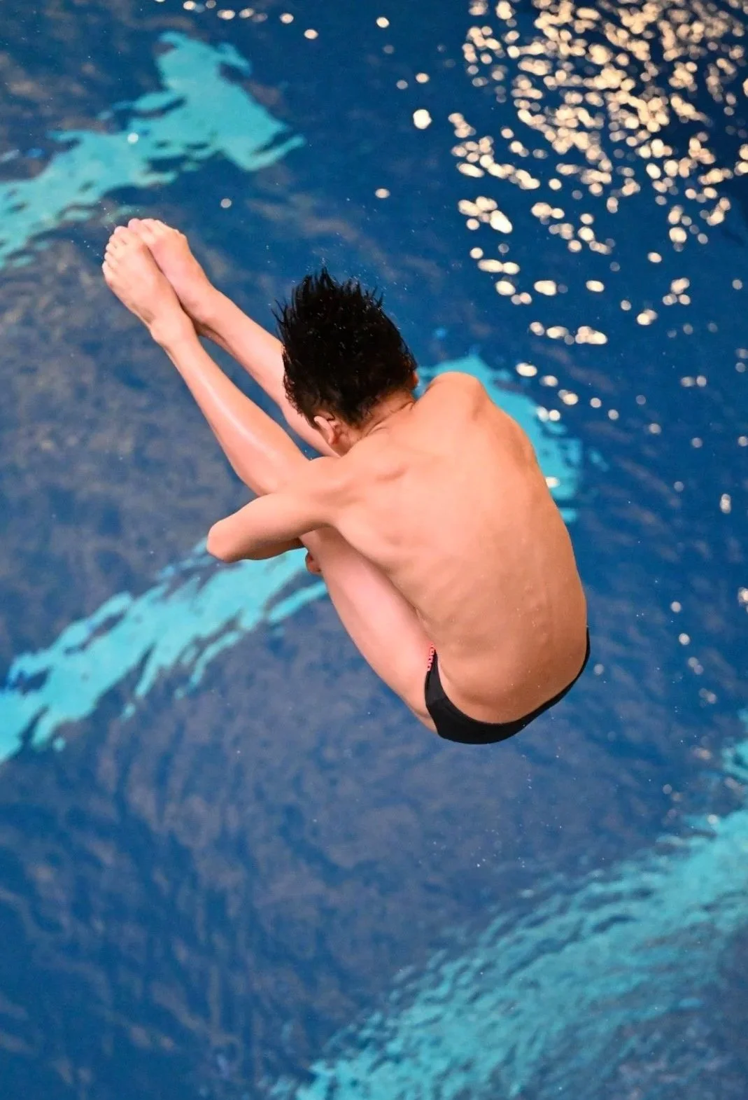A young male diver in black swim trunks is mid-air above a swimming pool, curlingly tucked with arms around his shins.