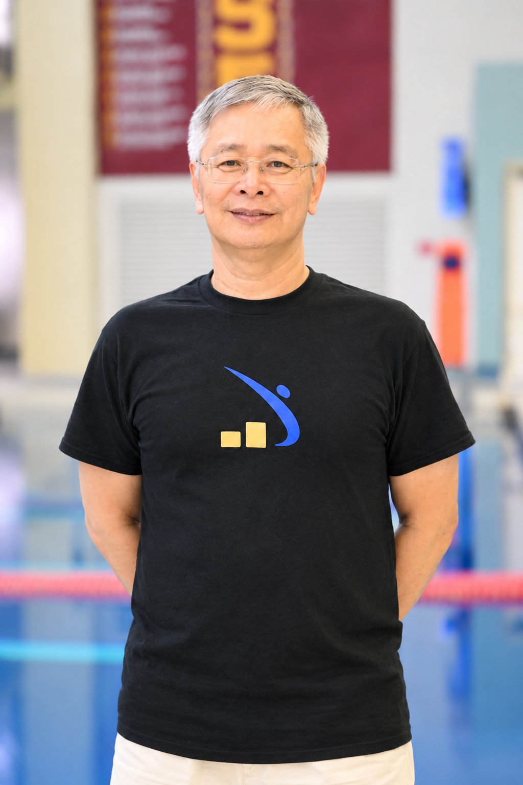 A smiling man with gray hair and glasses is standing indoors, wearing a black T-shirt with a blue and yellow logo on it.
