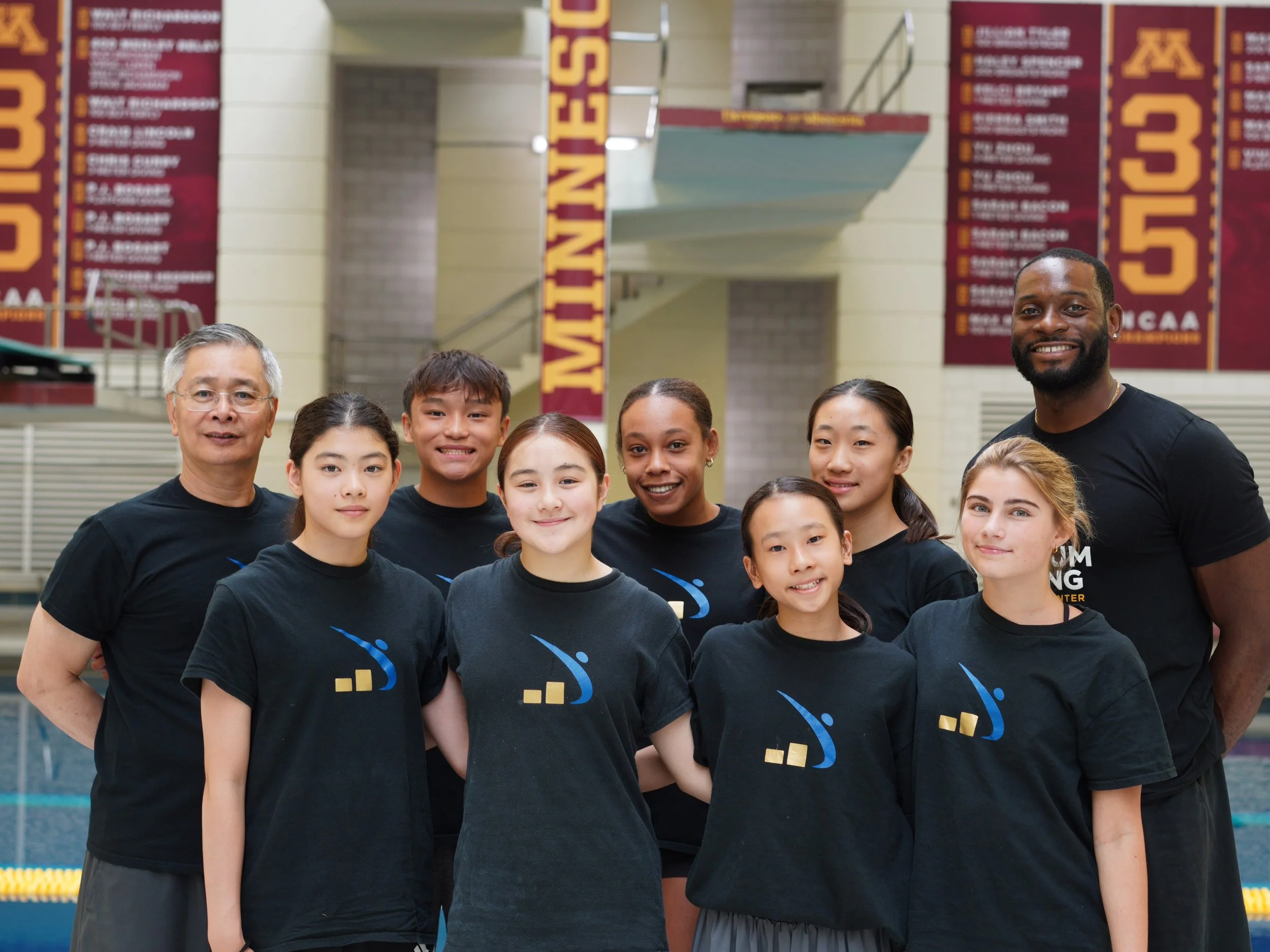 A group of young diverse divers and coaches Manny Pollard and Wenbo Chen posing in a swimming pool, with banners hanging in the background.