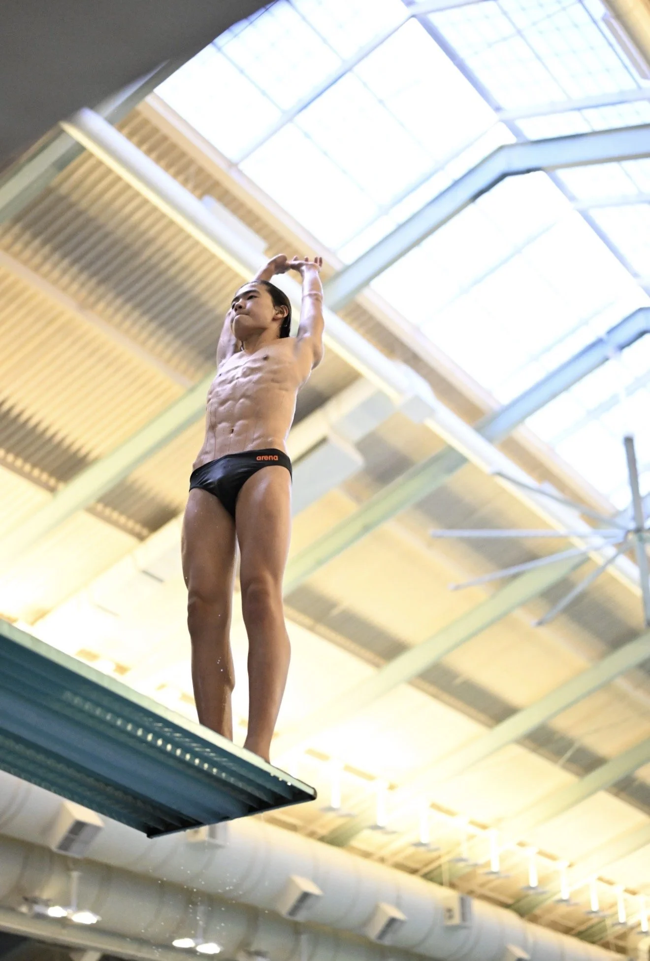 A male diver in black swim briefs standing on a diving platform at an indoor aquatic center, preparing for a dive under a glass ceiling.