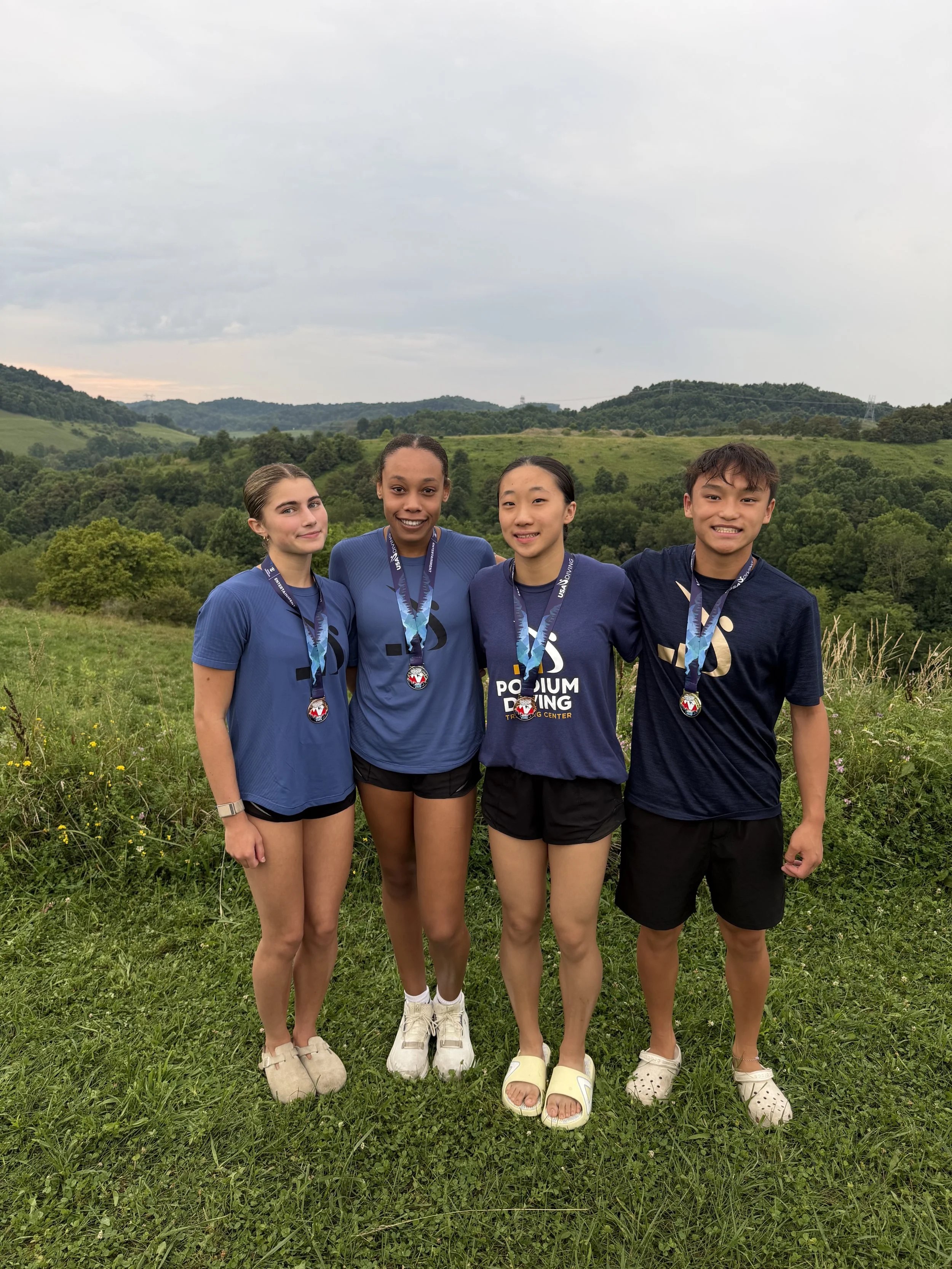 Four young diving athletes standing outdoors on grass, smiling, wearing athletic shirts and medals, with green hills and cloudy sky in the background.