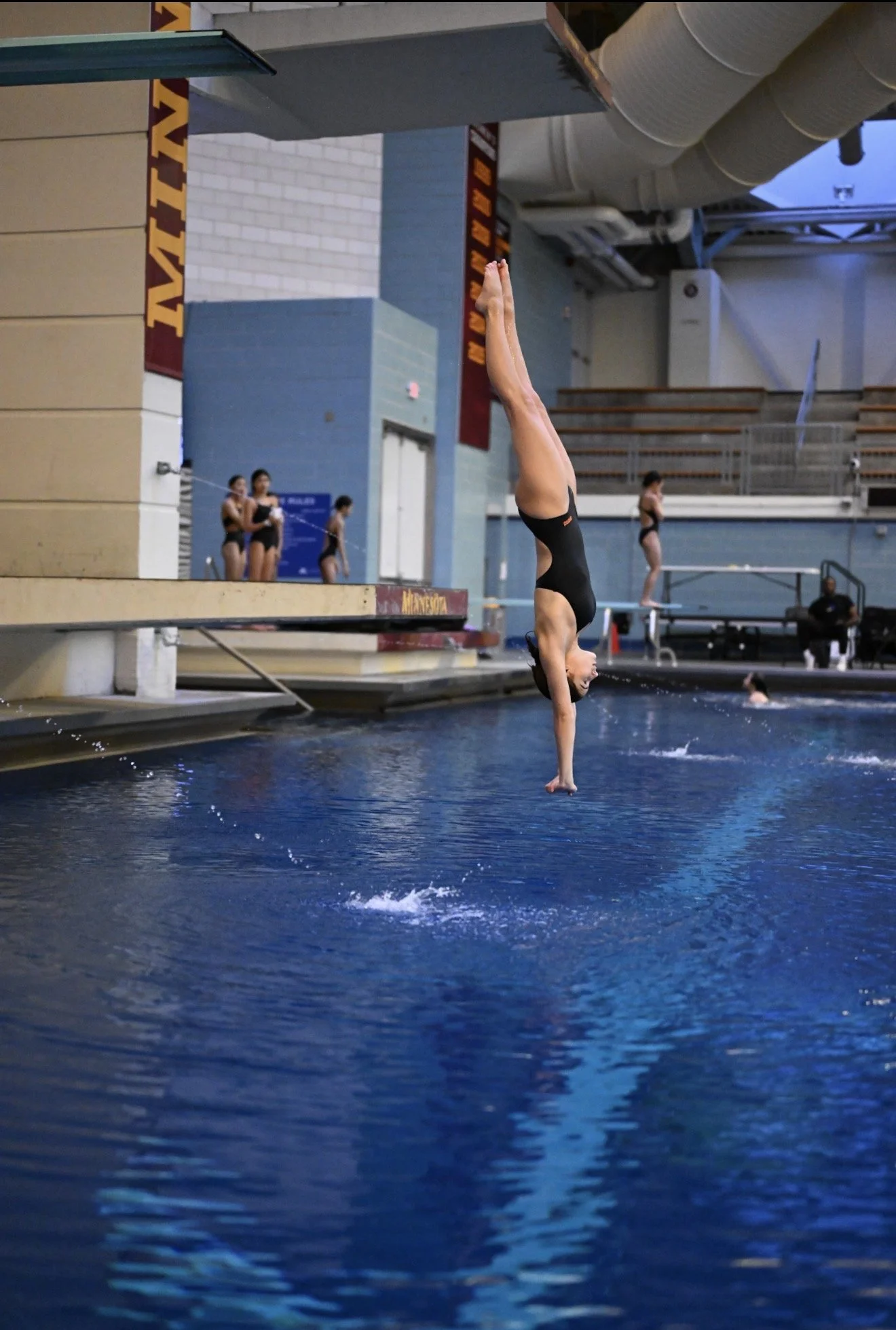 A female diver is executing a dive from a platform into a swimming pool during an indoor competition. There are three people standing on the side of the pool, and some others in the background.