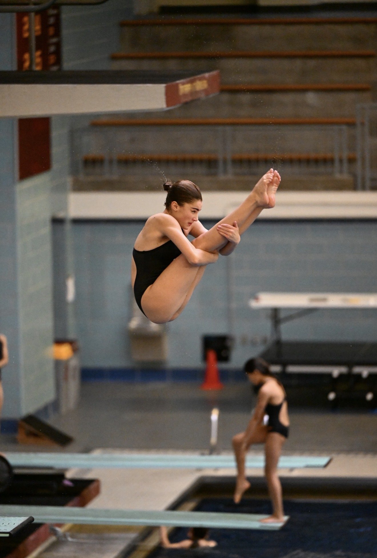 Female diver in a black swim suite performing a high-level pike dive move during an indoor diving training session or competition, with other divers visible in the background.