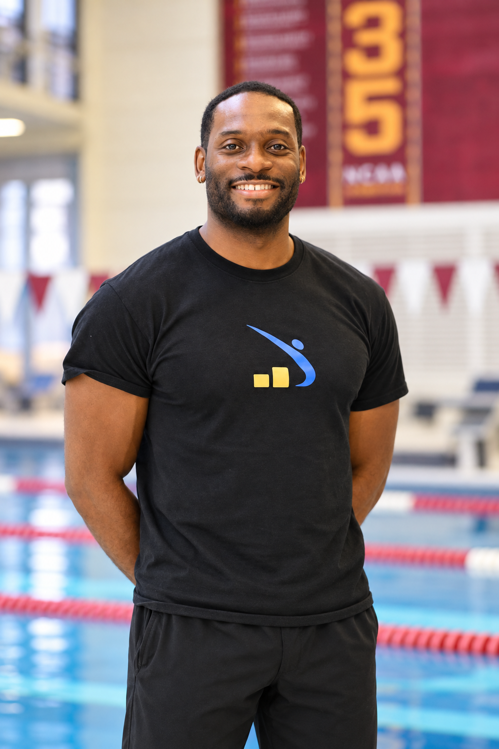 Coach Manny Pollard standing by an indoor swimming pool, wearing a black T-shirt with a blue and yellow logo, with a red banner in the background.