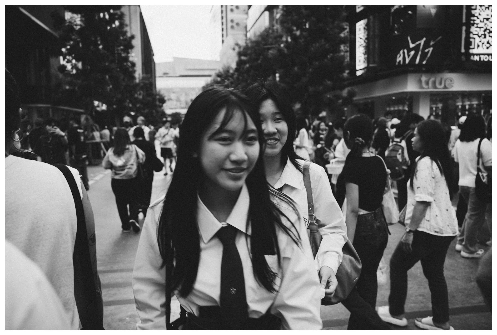 Two young women in school uniforms standing close together in a busy city street, surrounded by other pedestrians and buildings.