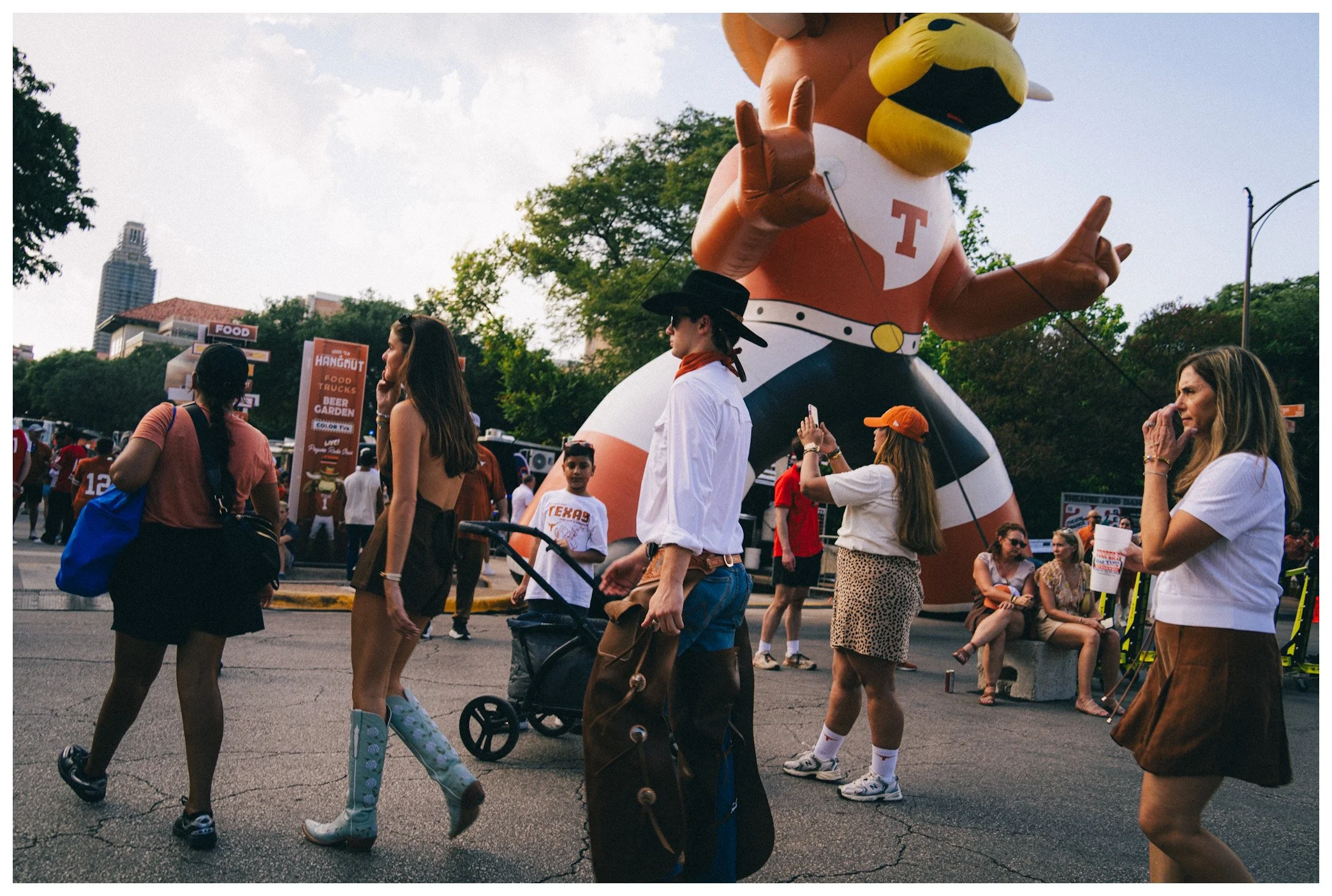 Crowd of people walking at an outdoor event with a large inflatable mascot in the background, featuring a yellow beak, glasses, a cowboy hat, and a white speech bubble with a red 'T' on it.