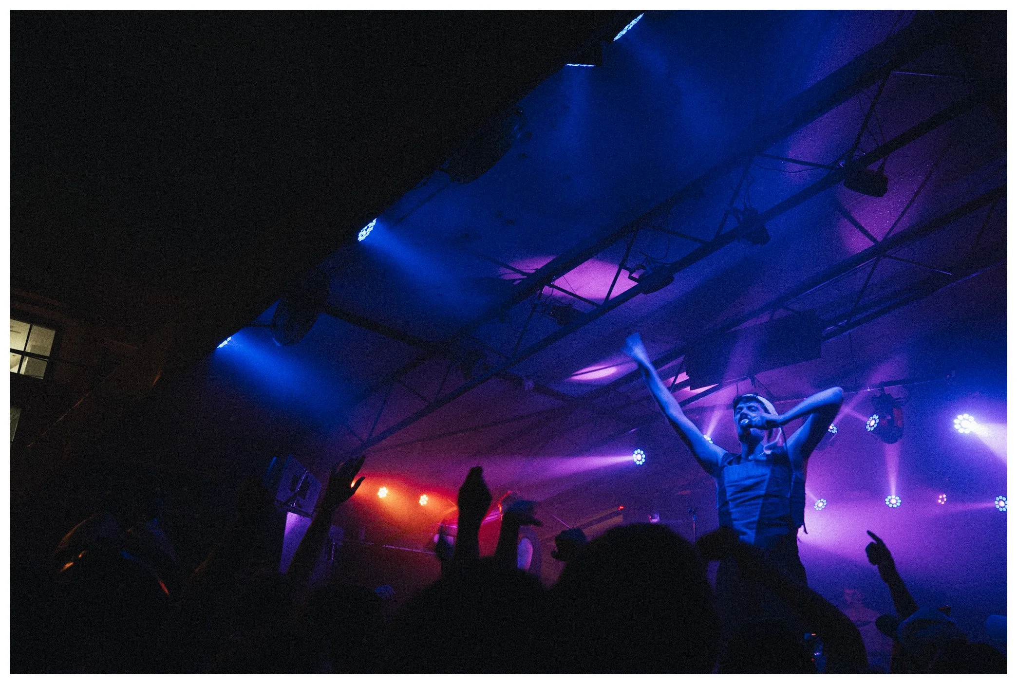 A performer with raised arms on stage under colorful blue, purple, and red lights, with audience members visible in the foreground.