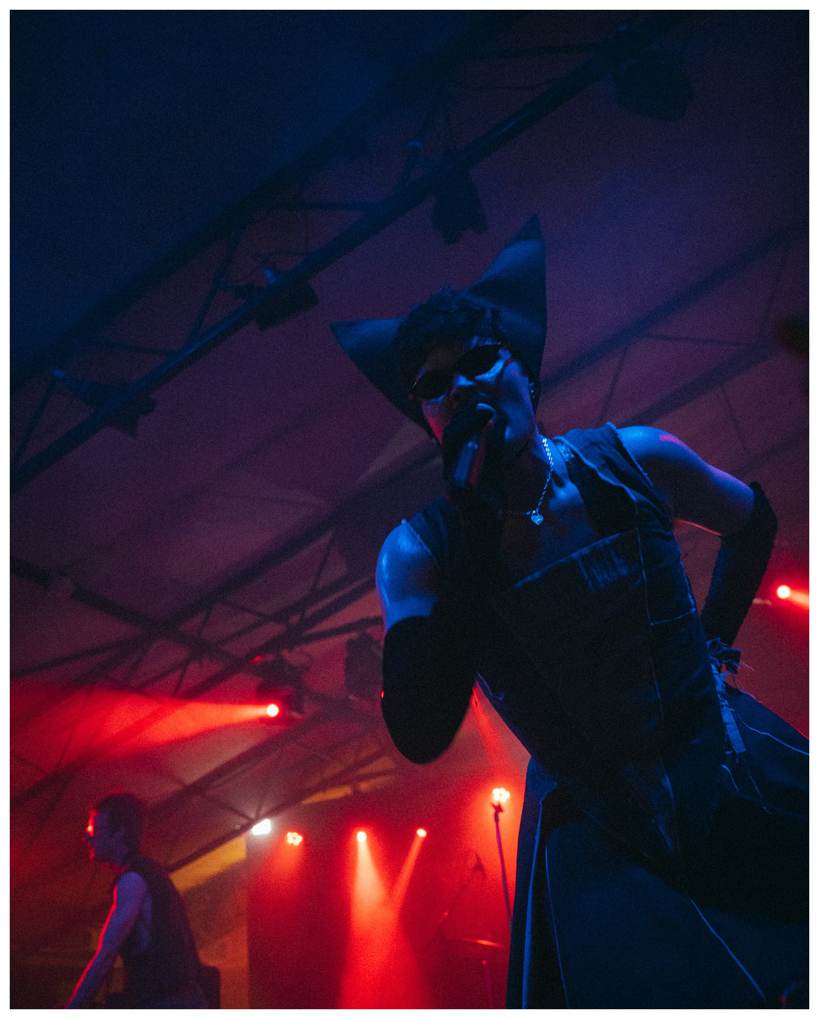 A performer with cat ears and sunglasses on stage under colorful red and blue lighting, possibly singing or speaking into a microphone in a concert or nightclub setting.