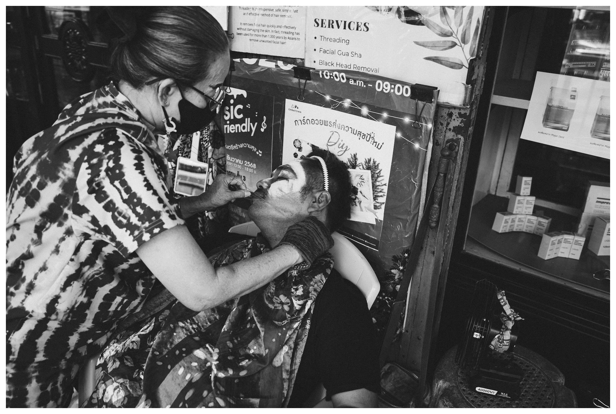 A woman with about-to-be facial makeup applying makeup to a man's face at a stall or shop. The woman is wearing a patterned top and a face mask, and the man has his eyes closed while sitting in a barber chair.