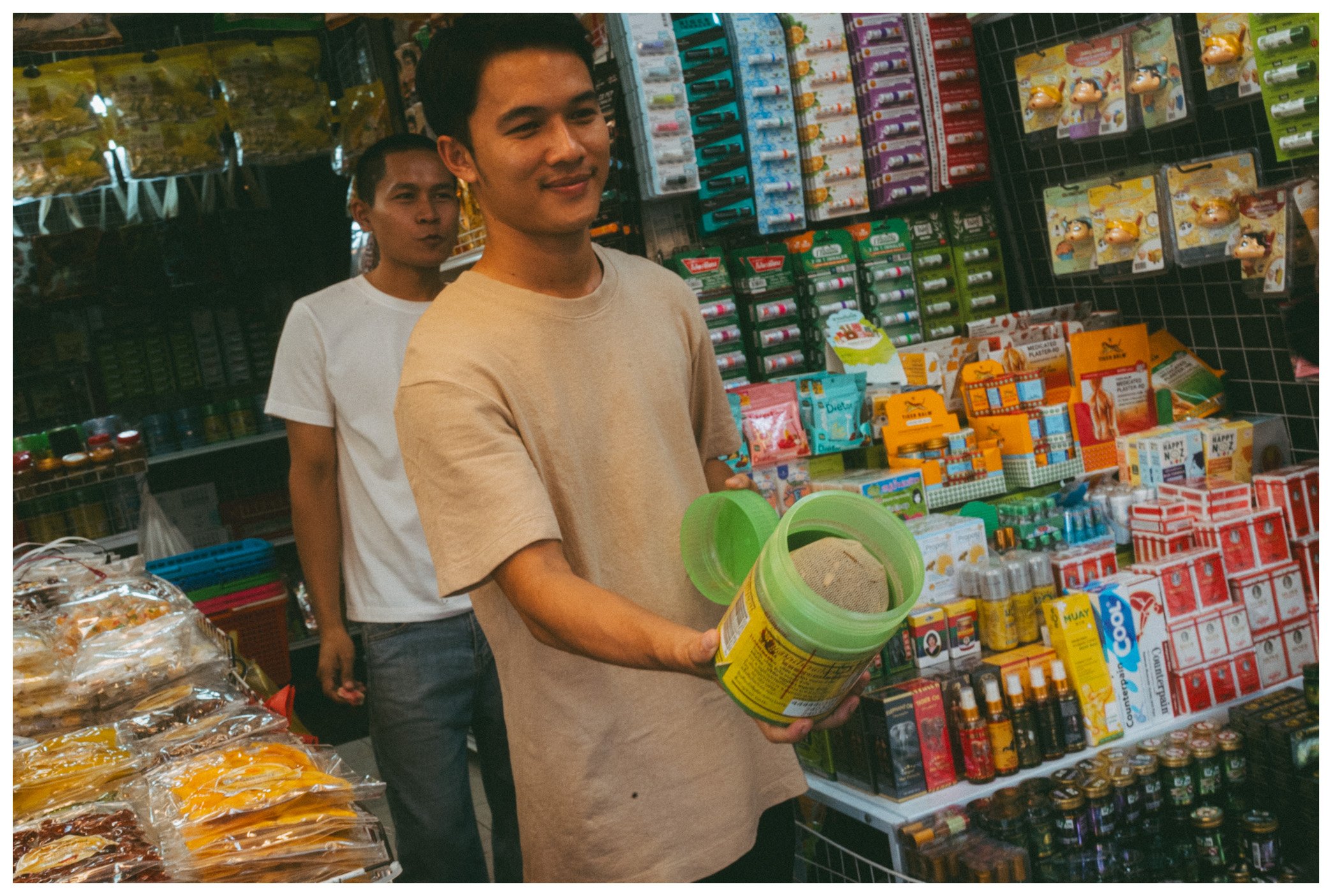 Two young men shopping in a store aisle with shelves stocked with various household and personal care products. The man in front is holding a plastic container and smiling, while the man behind him appears to be making a playful expression.