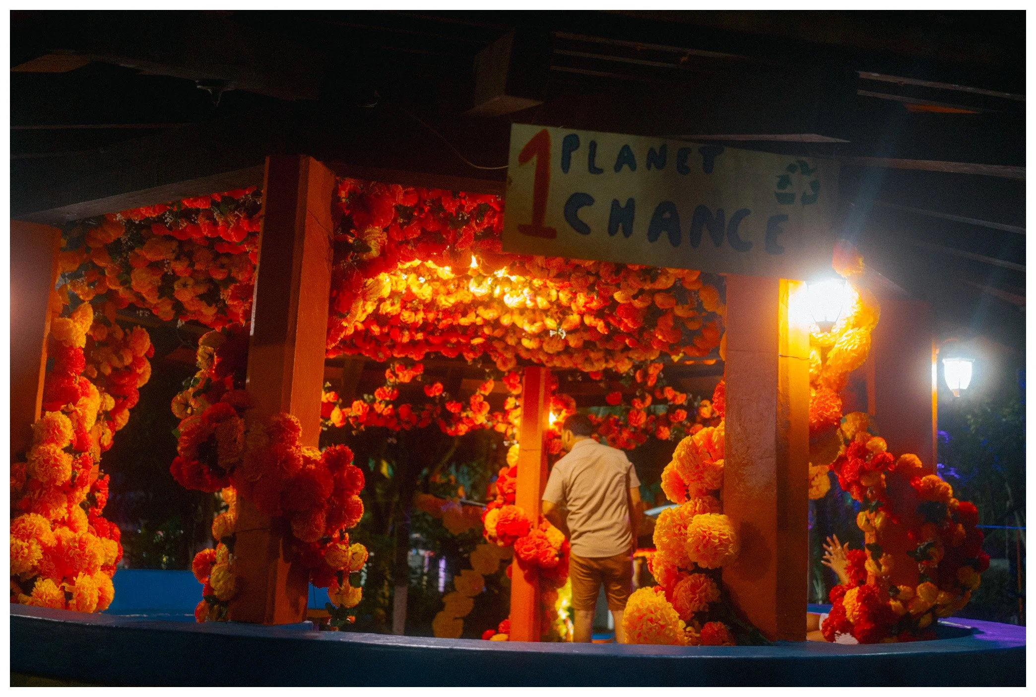 Nighttime scene of a floral entrance adorned with orange and yellow marigold flowers, with a sign that says '1st Planet Chance' and a person walking through the decorated area.