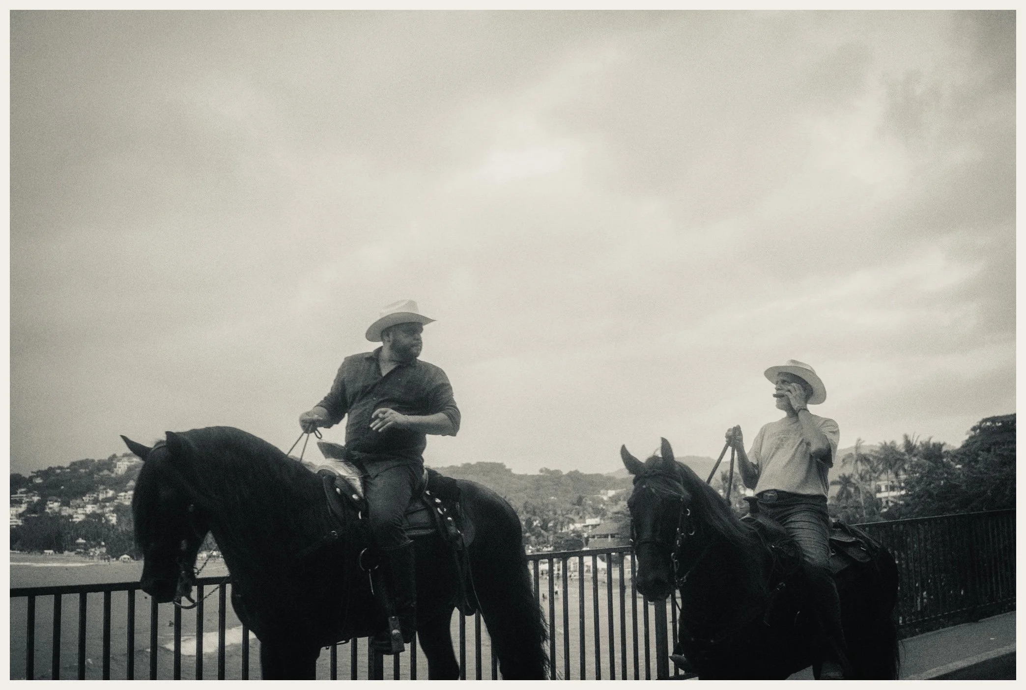 Two people riding horses on a bridge, wearing cowboy hats, with a cityscape and hills in the background.