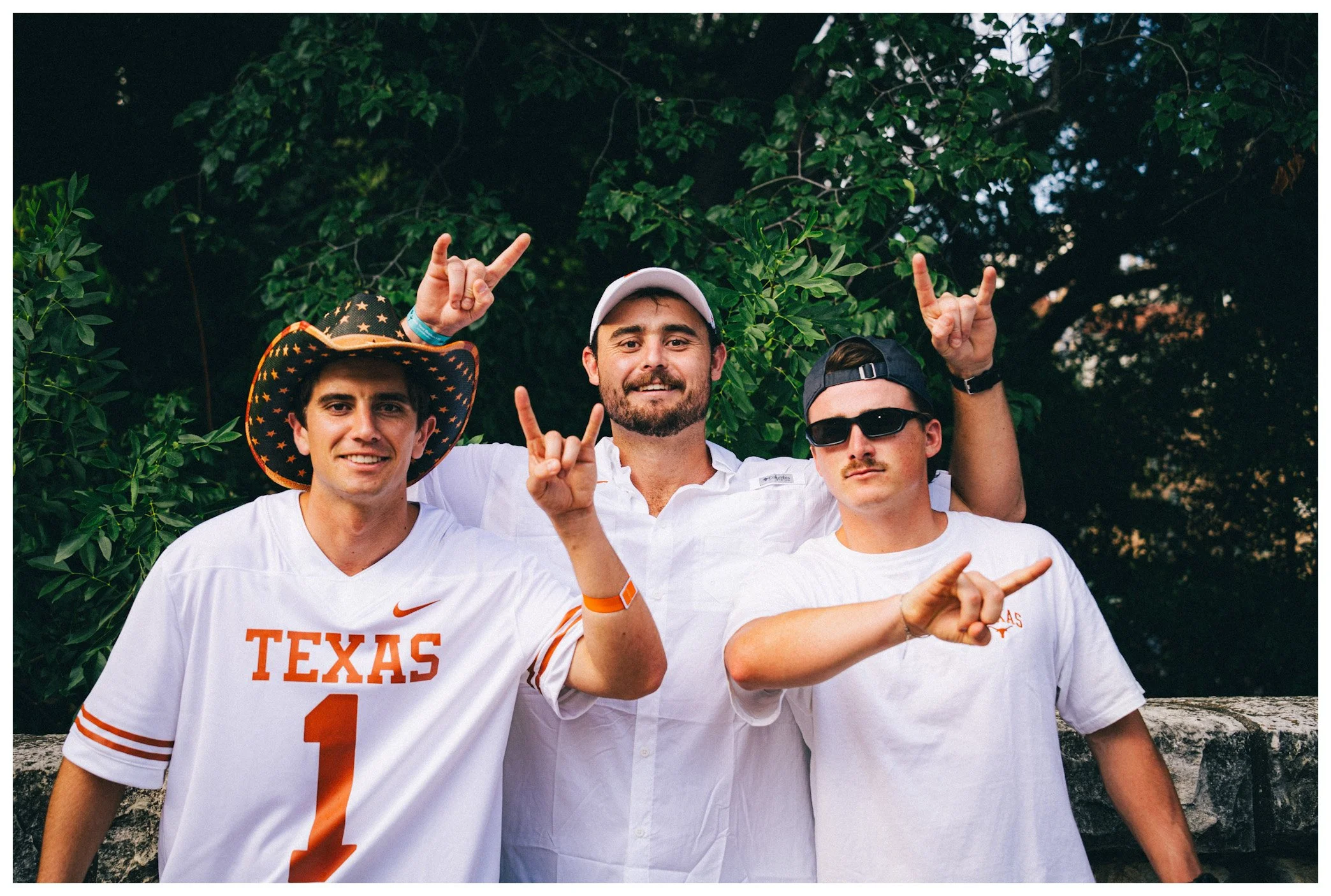 Group of four young men posing outdoors with lush green trees in the background, two wearing Texas Longhorns football jerseys, one in a cowboy hat, and all making hand gestures.