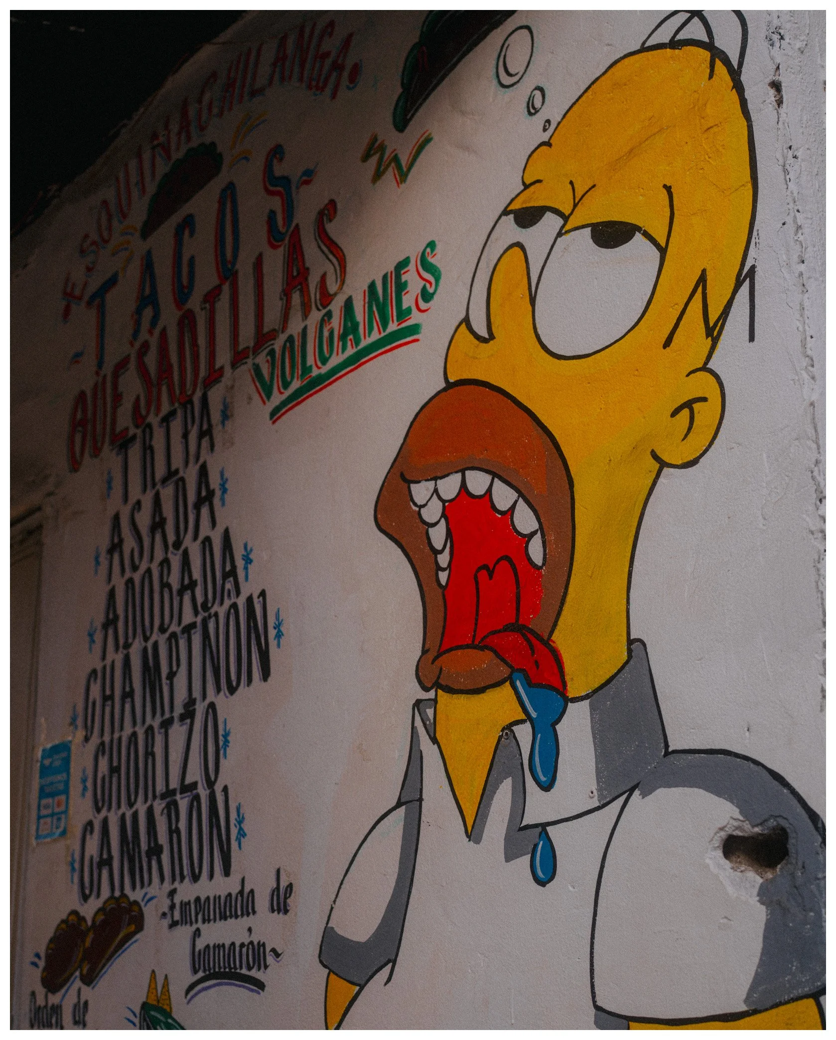 Street art painting of Homer Simpson with tears and a sad expression, with text in Spanish around him mentioning a story about salty fish, volcanoes, and chamorron.