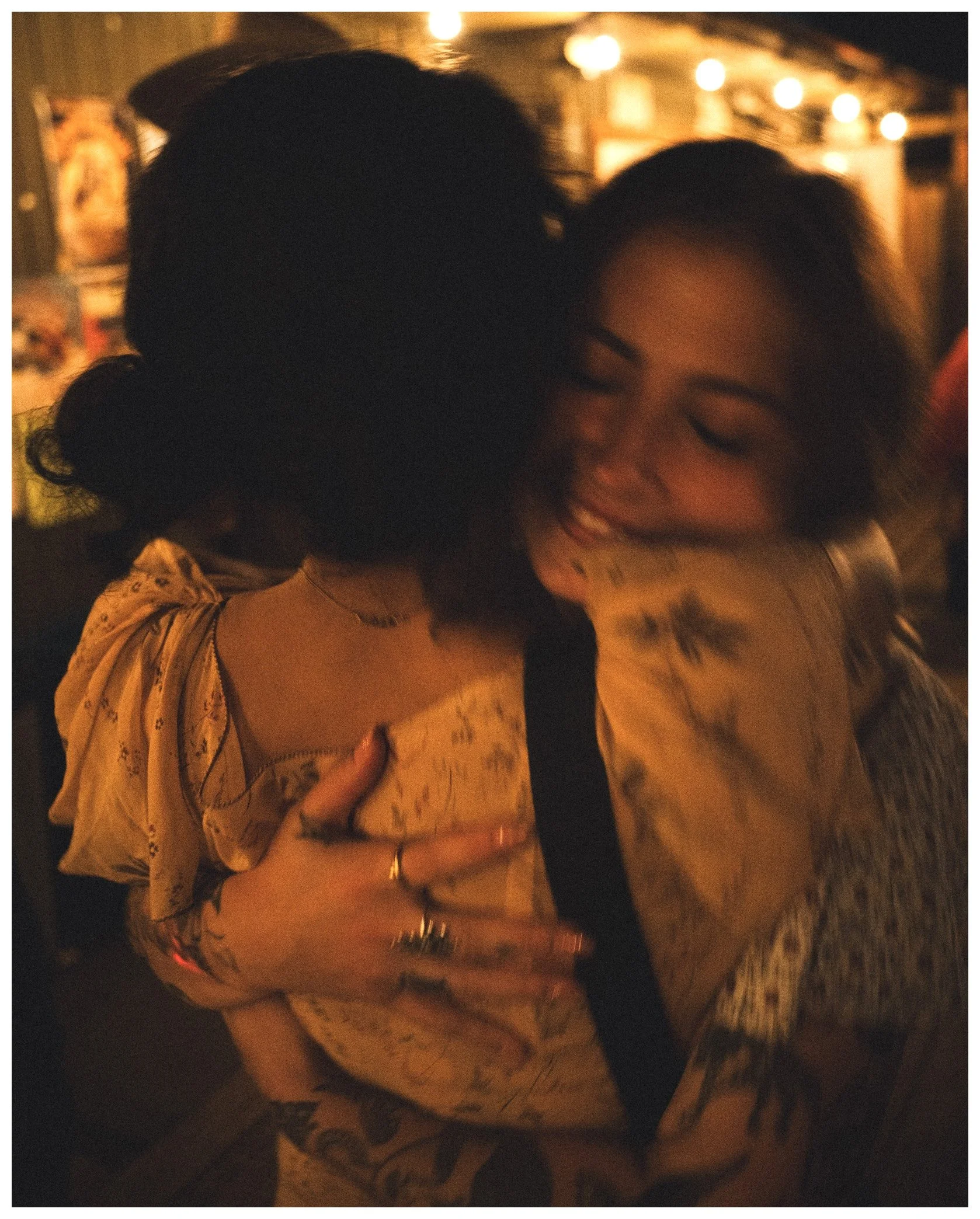 Two women hugging at a dimly lit party or social gathering with warm lighting and string lights in the background.