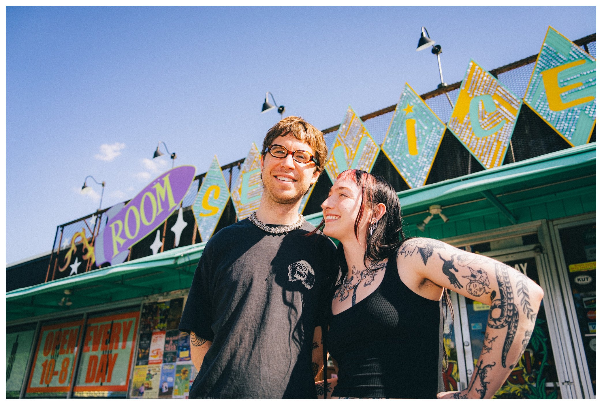 A young man with glasses and a young woman with tattoos smiling together outside a colorful amusement park or carnival, with signs that say 'BOOM,' 'SILVER,' and 'OPEN EVERY DAY 10-8.'