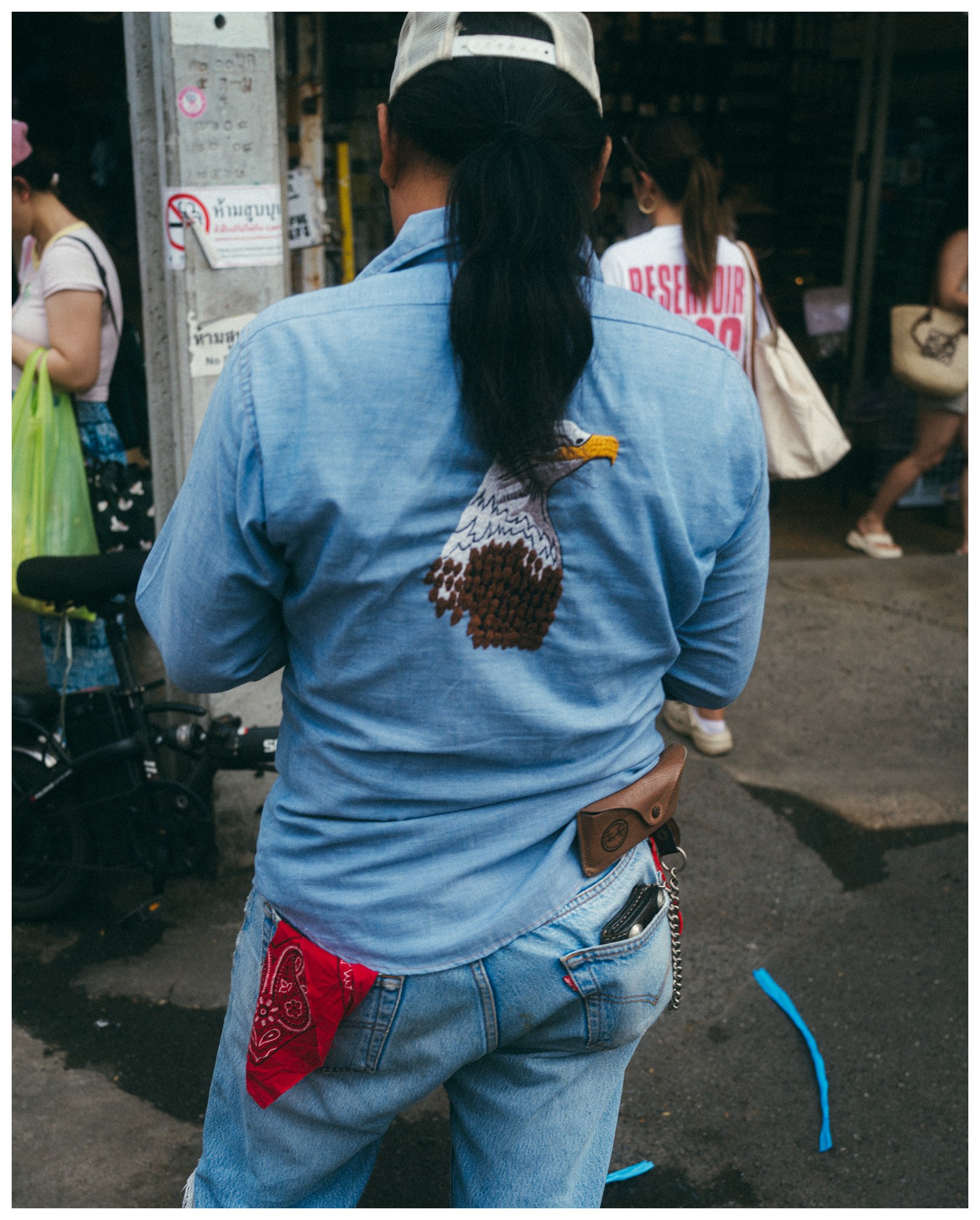 Back of a person wearing a light blue denim jacket with an embroidered eagle on the back. The person has long dark hair tied in a ponytail and is standing outdoors near several other people and a motorcycle.