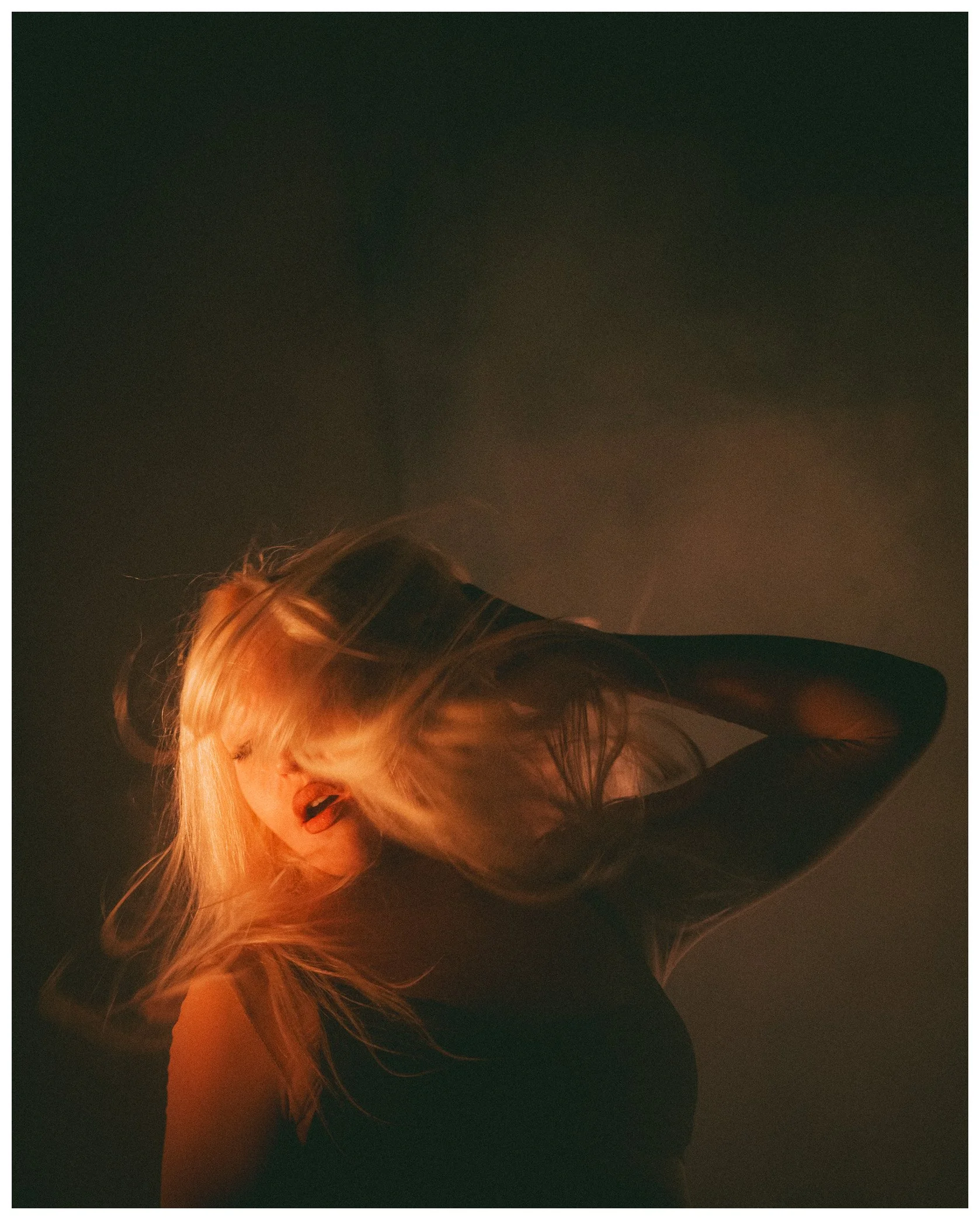 A woman with long blonde hair and a black top, with her head tilted and looking down, in a dimly lit environment with a dark background.