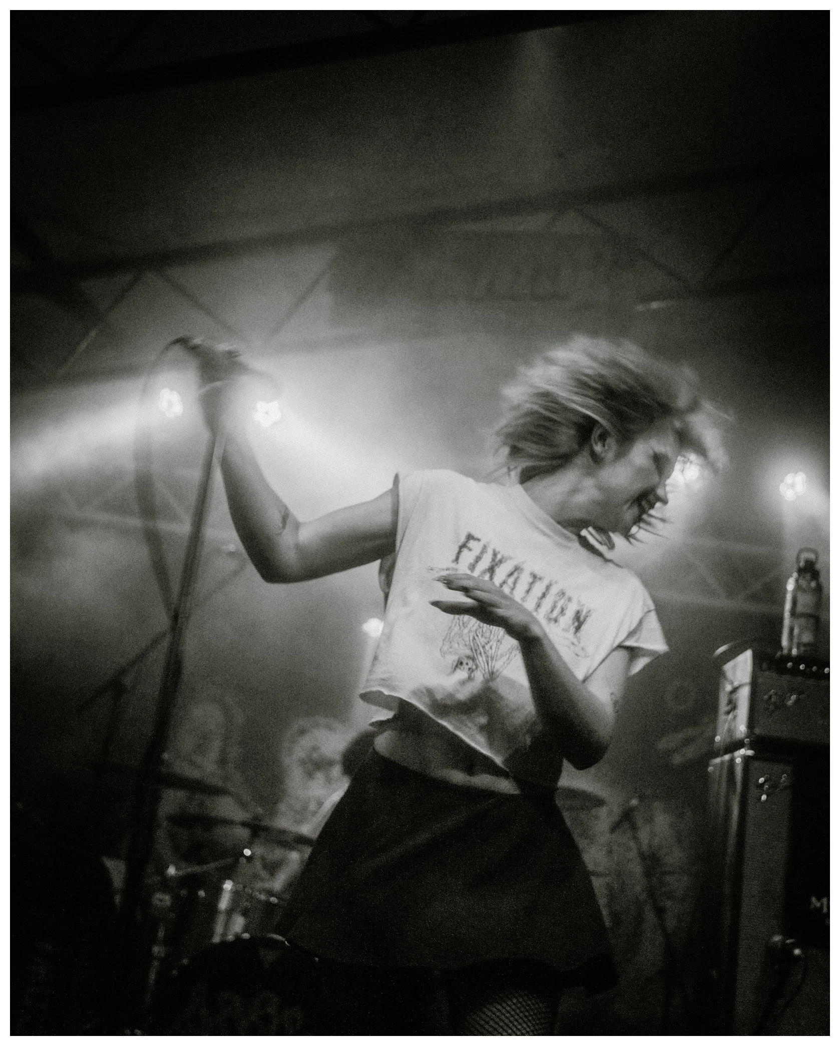 Person riding a motorcycle or scooter at night, illuminated by bright lights, with hair blowing and wearing a casual t-shirt.