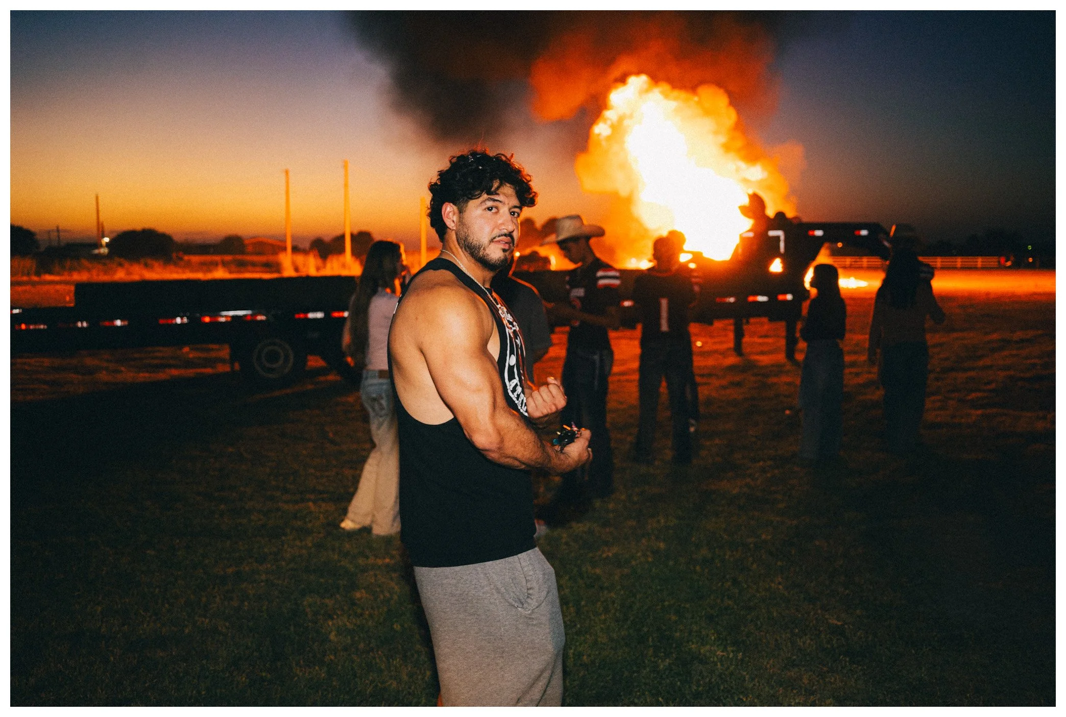 A young man with curly hair and a beard, wearing a sleeveless shirt and gray sweatpants, standing with his arms crossed in front of a fiery explosion in the background at dusk, with a group of people and trucks nearby.