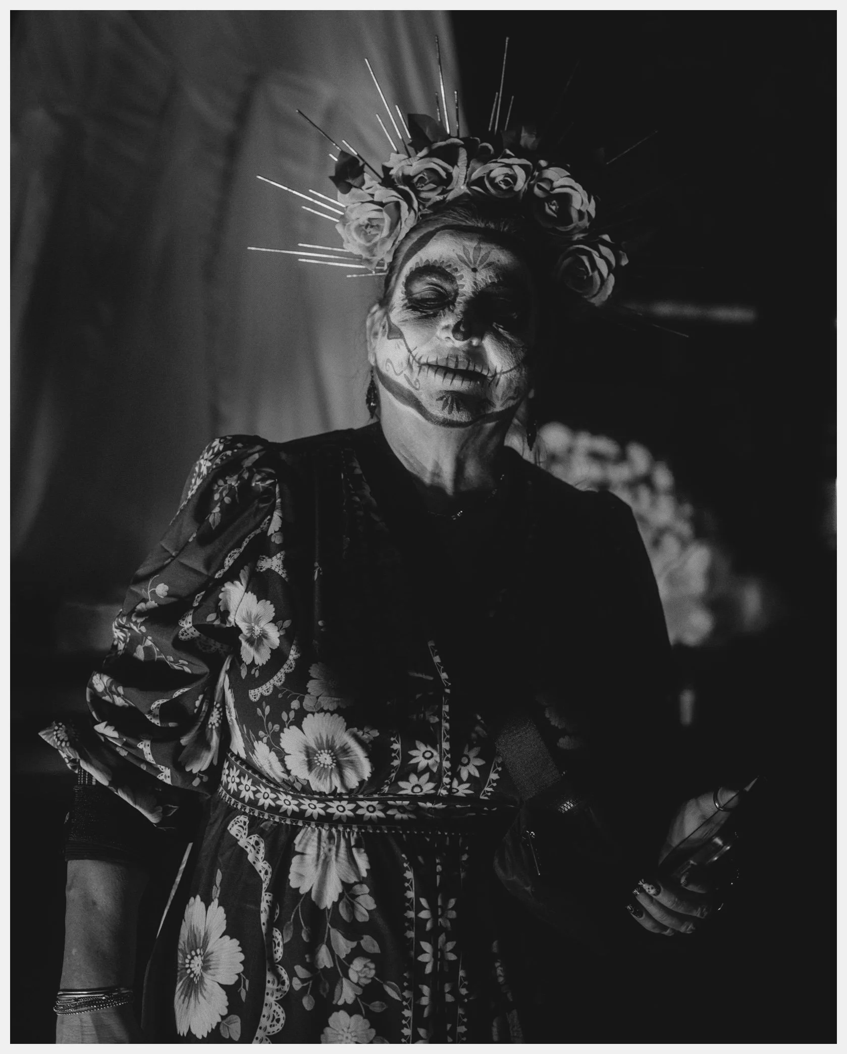 A person wearing traditional Mexican sugar skull face paint, adorned with face paint details, a floral headpiece with roses, and a patterned blouse, holding a wine glass, in a black and white photograph.