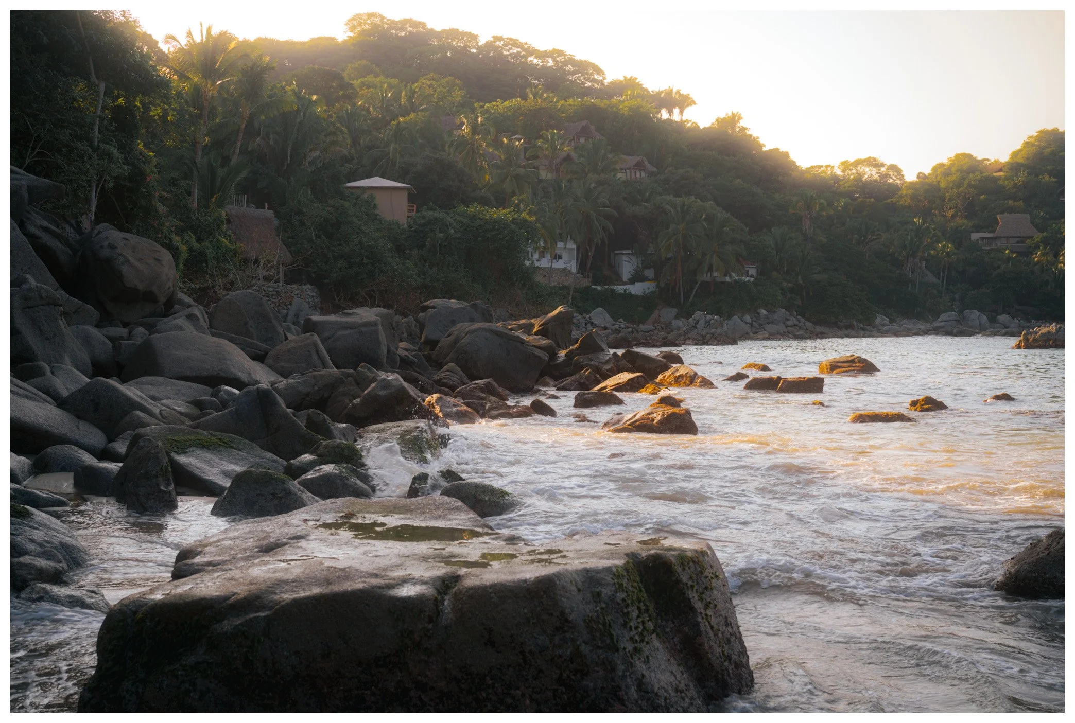 Rocks along the shoreline with water in the foreground and lush hillside with trees and houses in the background, during sunset.