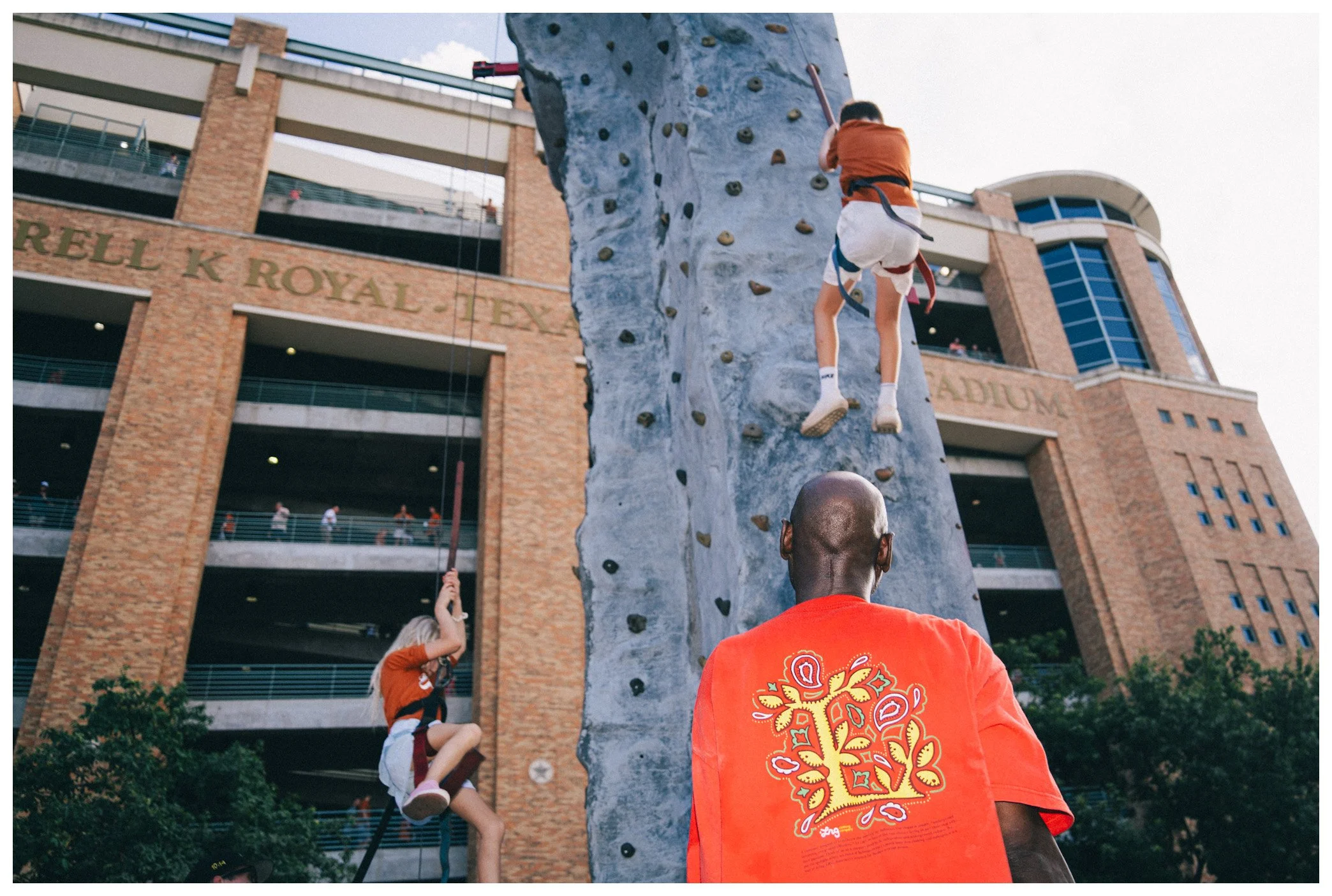 People climbing an outdoor wall at a stadium with brick and glass architecture, some spectators watching from balconies.