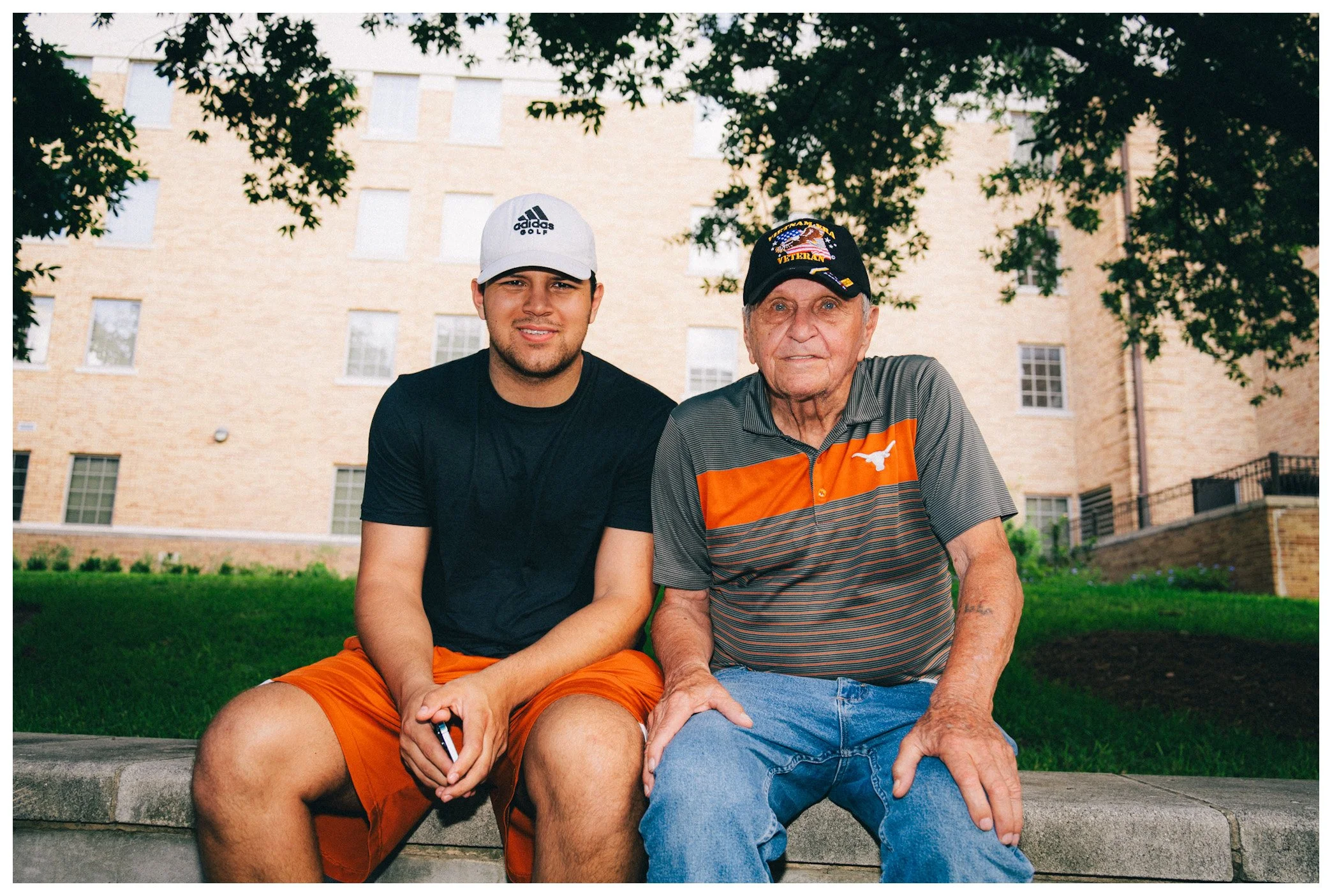 A young man and an elderly man sitting outdoors on a park bench, smiling. The background features a brick building with windows and shaded trees.