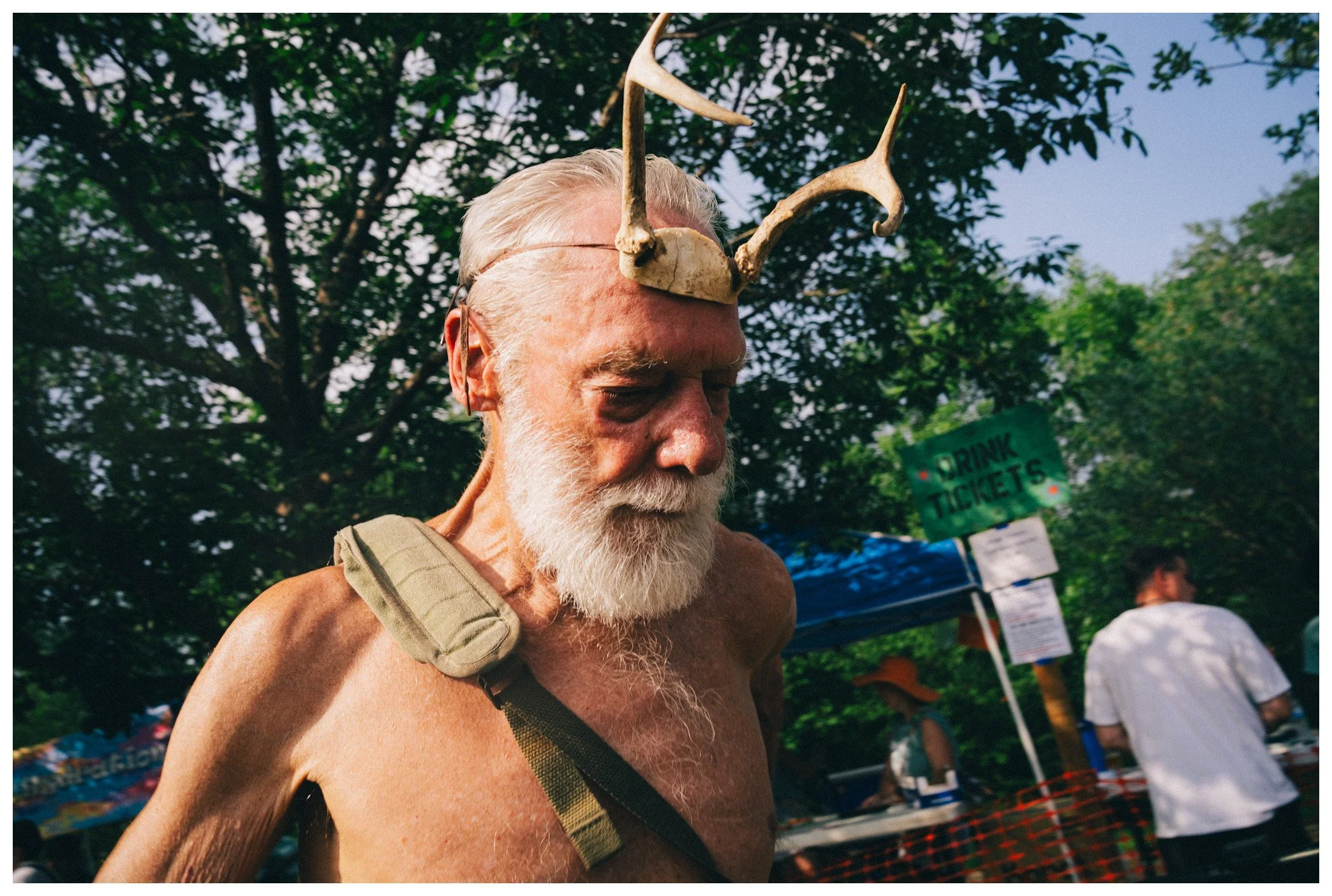 An older man with white hair and beard, shirtless, wearing a pair of sunglasses on his head and a strap around his neck, has an animal skull with antlers balanced on his head. He has a serious expression and is outdoors with trees and people in the b