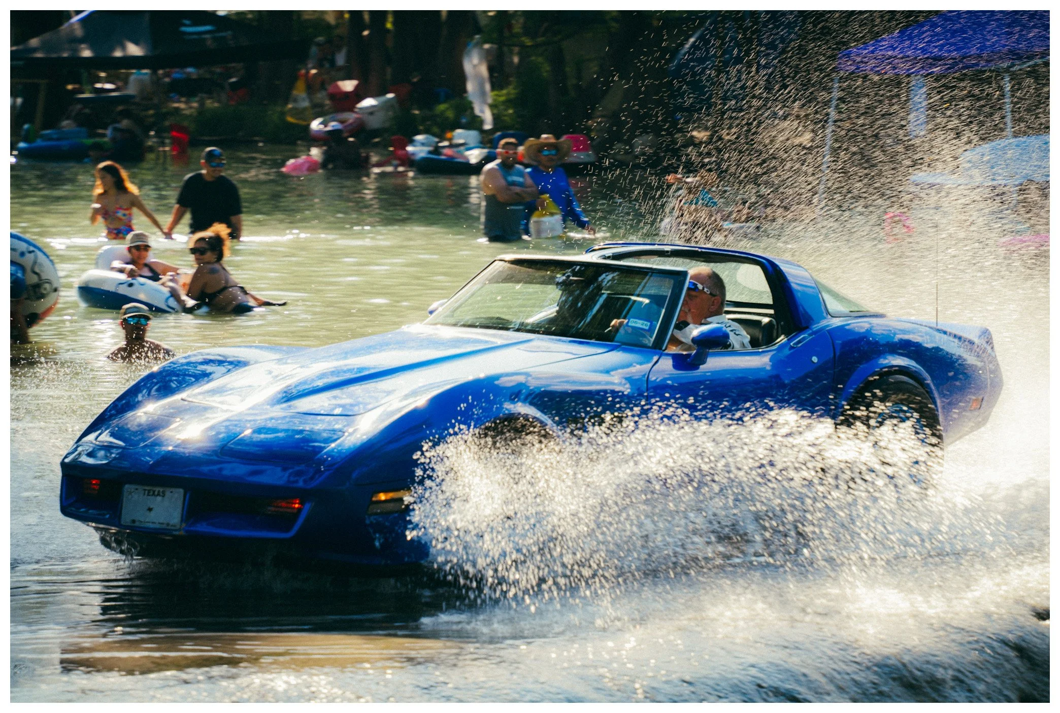 A blue sports car driving through water, creating a splash, with people in the water and on the shore in the background during daytime.