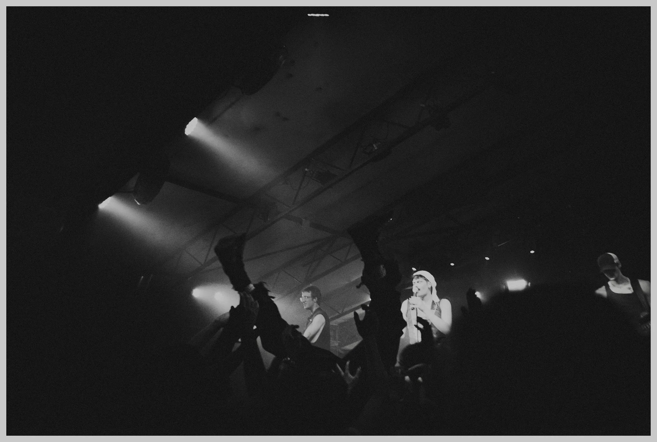 A group of people in a dimly lit club or concert venue, with spotlights overhead, some people are standing and talking, and one person is holding a drink.