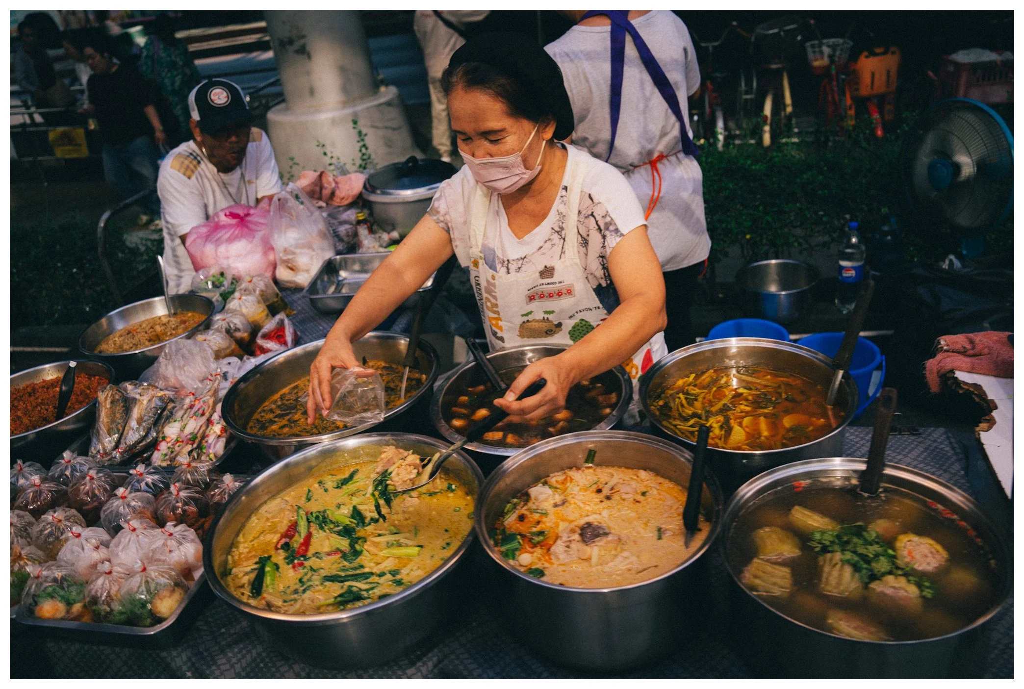 A woman in a face mask preparing food at a street food stall filled with large pots of various soups and dishes, with other people and cooking equipment in the background.