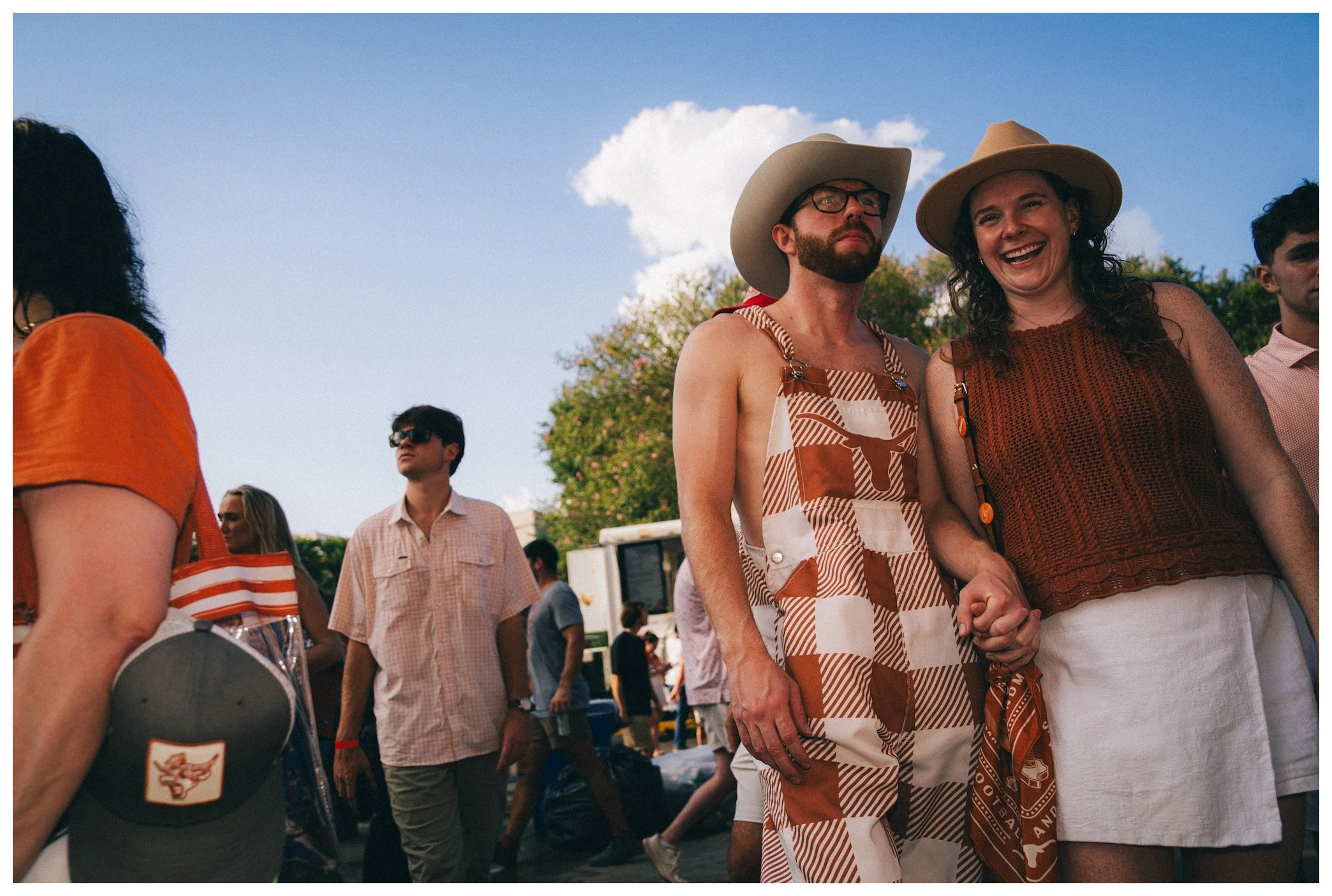 People attending an outdoor event, likely a festival, with a woman smiling and holding hands with a man wearing a cowboy hat.