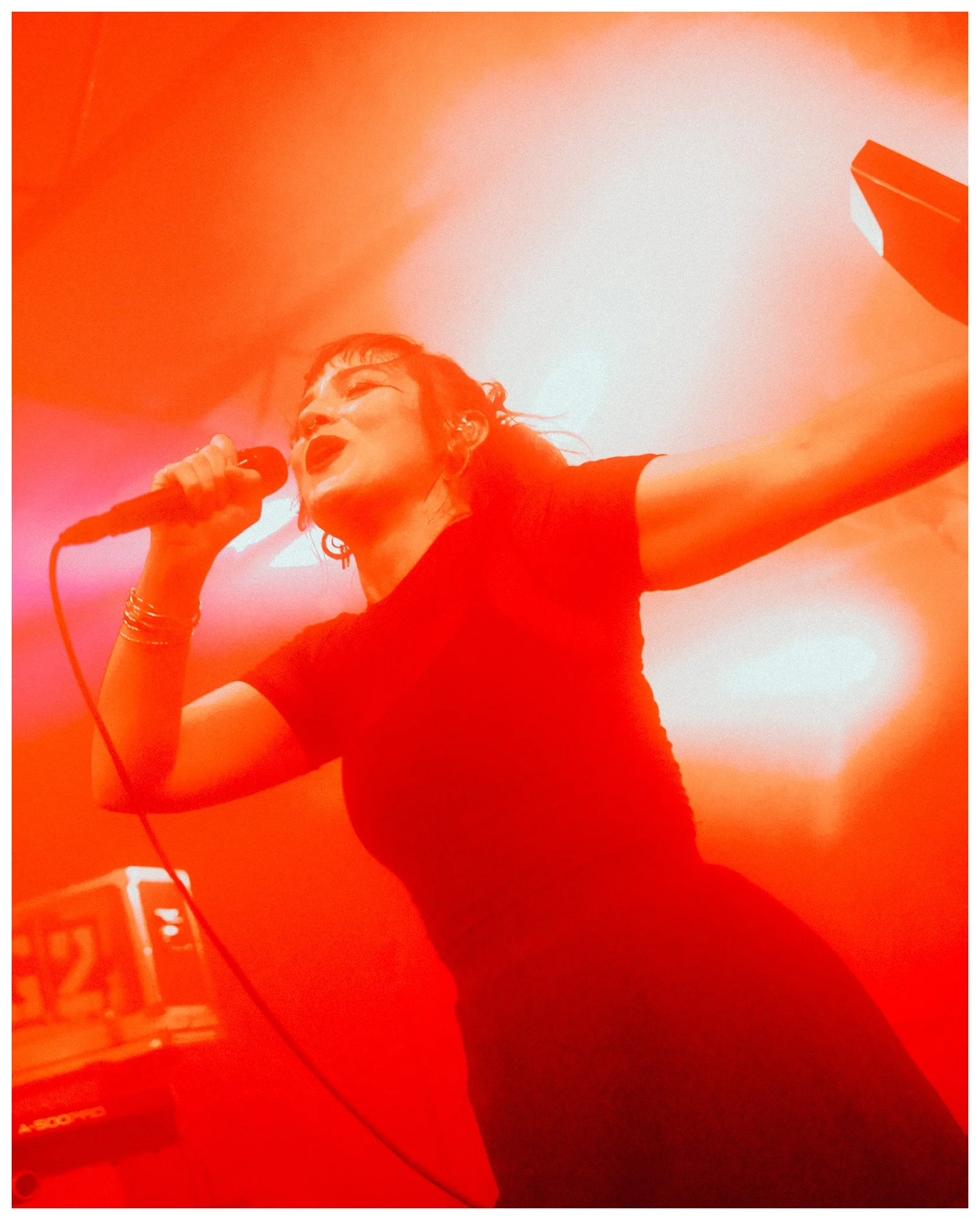 A woman singing into a microphone on stage, with her eyes closed, wearing earrings and bracelets, and an orange background.