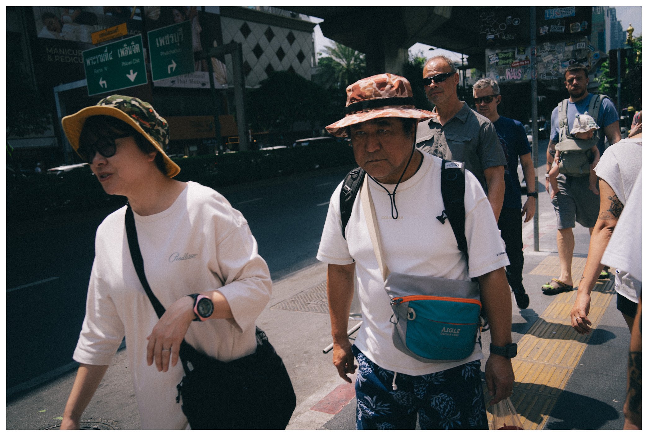 Group of pedestrians walking on a city sidewalk under an overpass, including an older woman with sunglasses and a man in a hat and sunglasses in the foreground, with various signs and graffiti in the background.