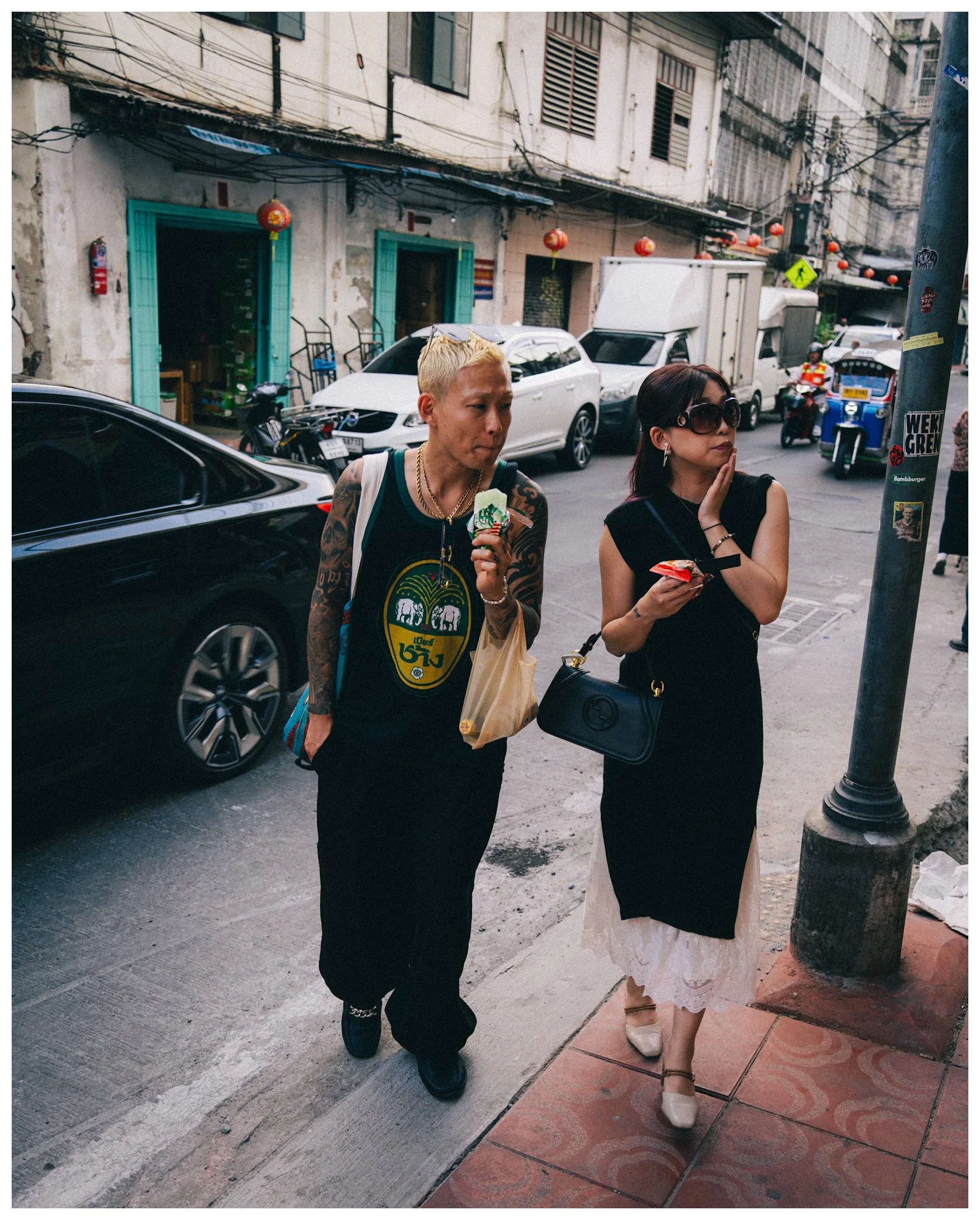 Two women walking on a sidewalk in an urban area with vehicles and buildings in the background, decorated with red lanterns.